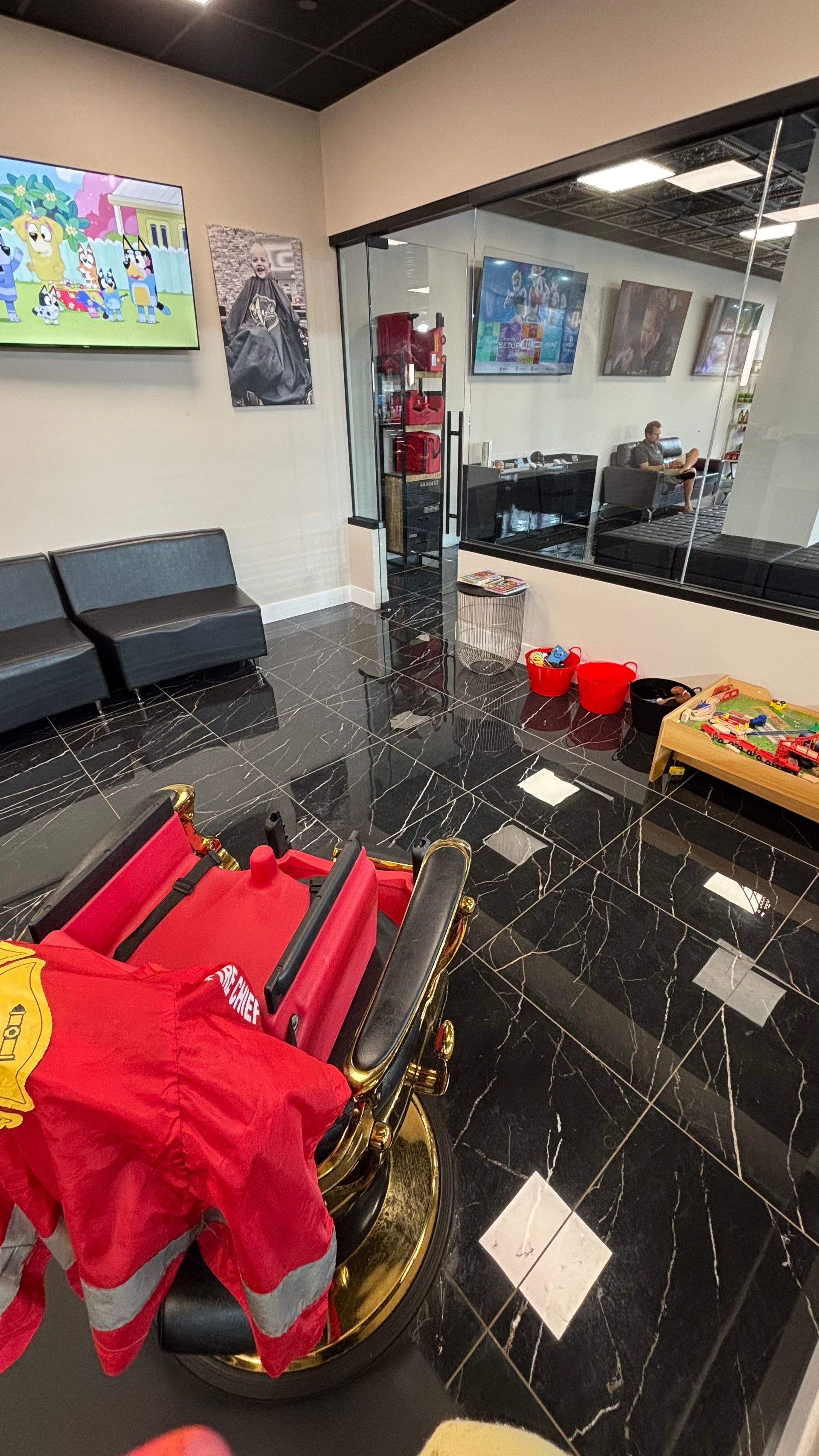 Barber shop interior with black tile floor, waiting chairs, wall art, mirrors, and red salon equipment.
