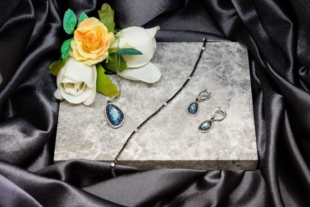 Jewelry set with blue teardrop-shaped gems on a gray stone, with flowers and black satin background.