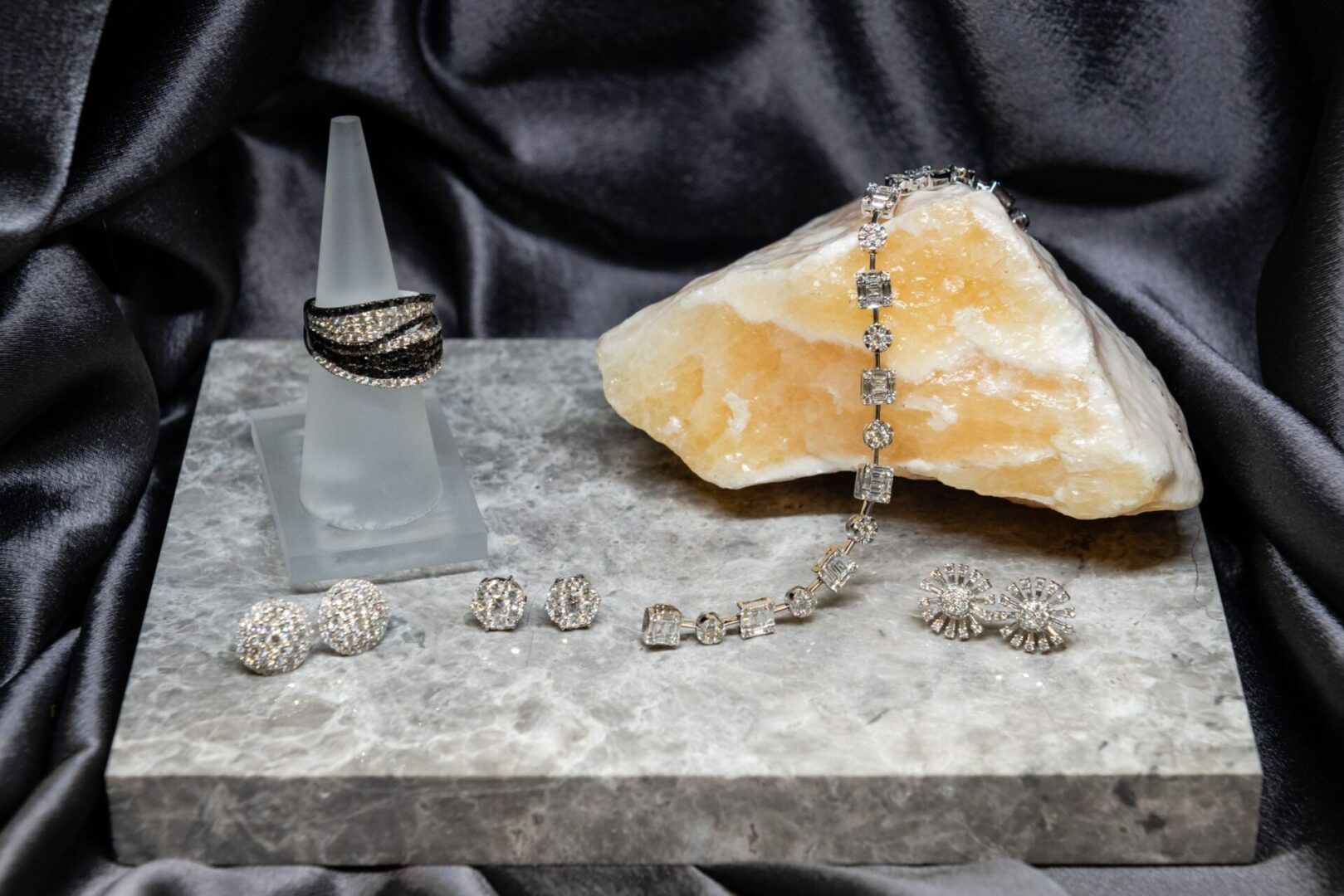 Jewelry display: black and silver ring on a stand, bracelet on a rock, earrings, set on a gray surface with dark gray fabric.