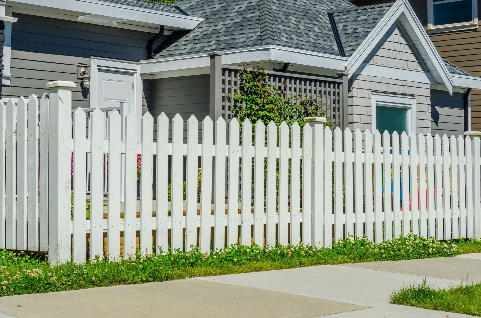 White picket fence in front of a gray house with a dark gray roof. Green grass and sidewalk in front.