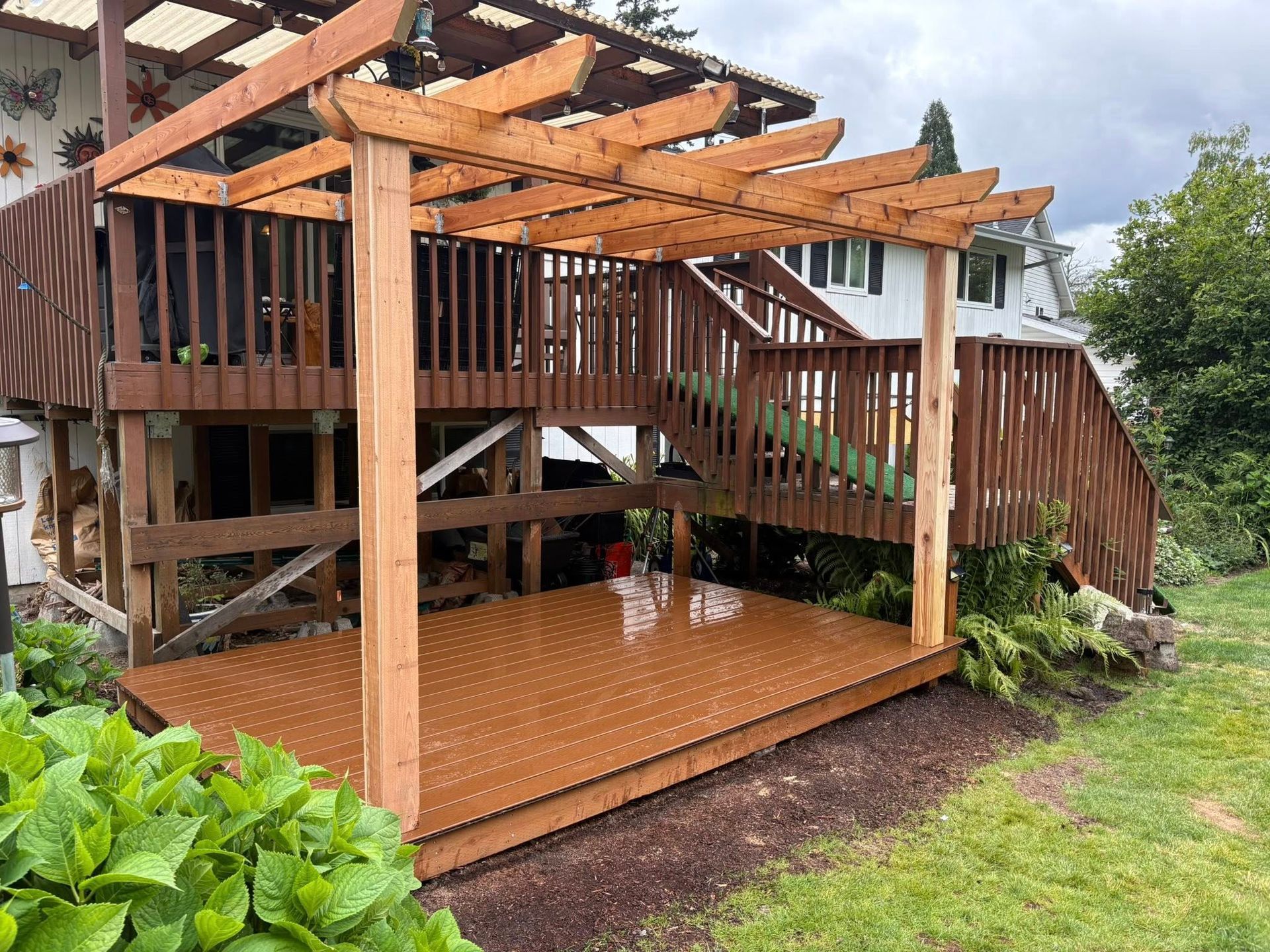 A wooden pergola sits over a newly stained, dark brown patio deck built beneath an existing raised wooden deck.