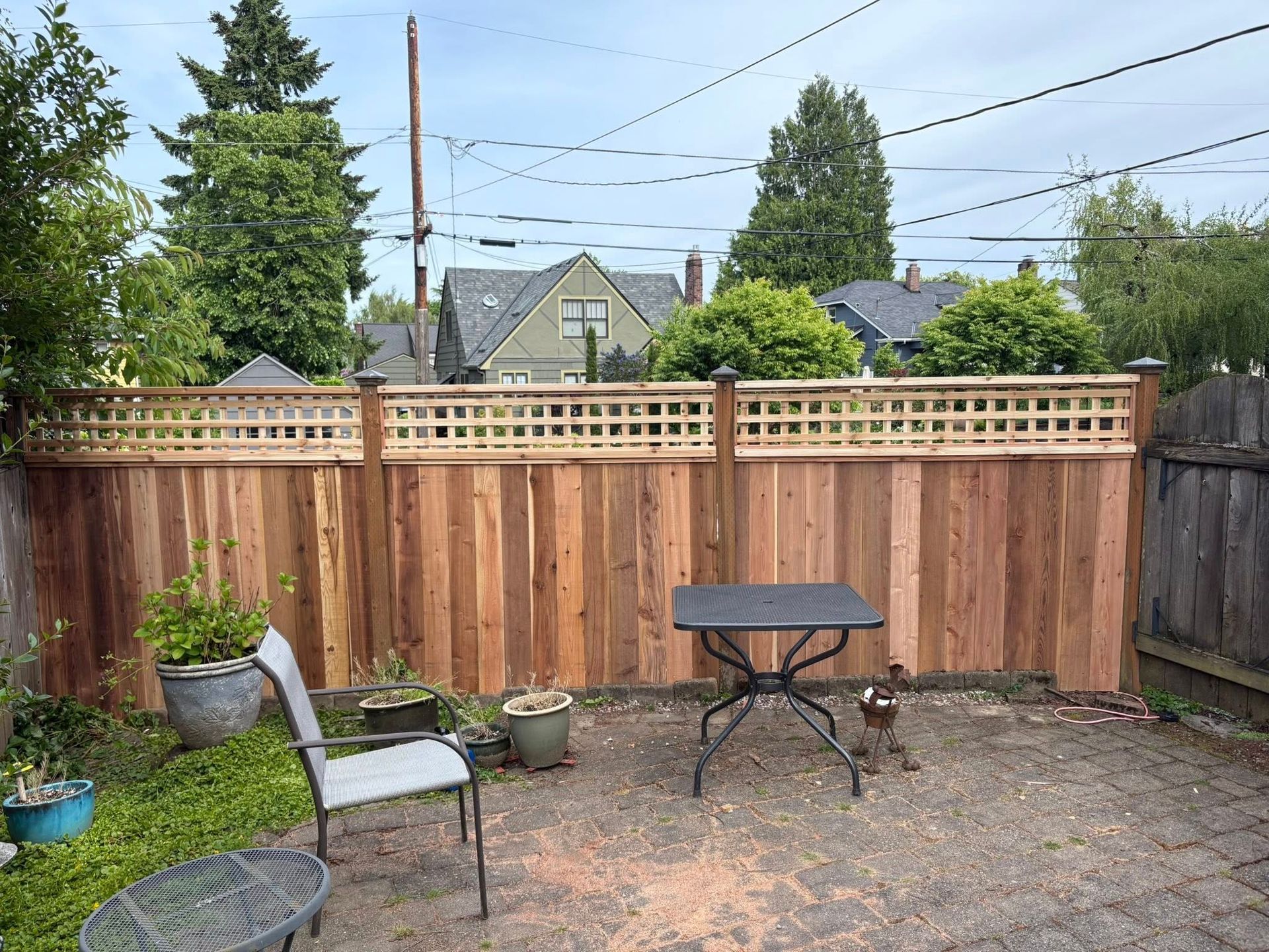 A backyard patio featuring a new cedar wood fence with a lattice top, a small table, a chair, and potted plants.