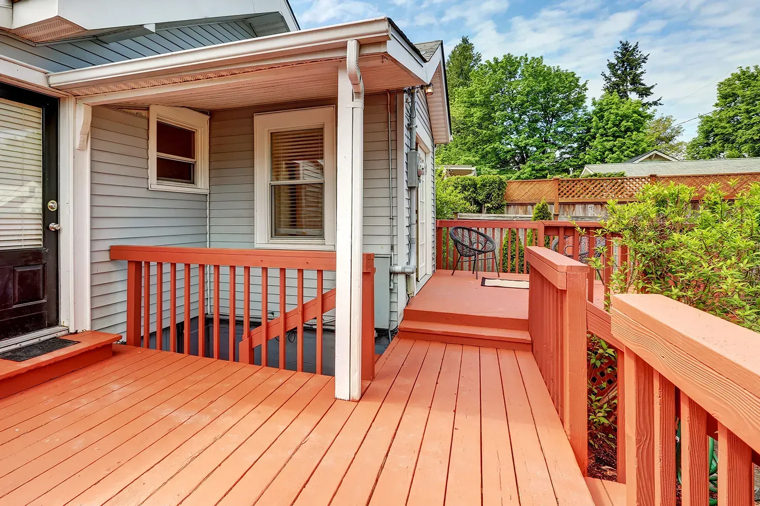 Wooden deck and railing attached to a light blue house. Orange deck with steps. Green trees in the background.