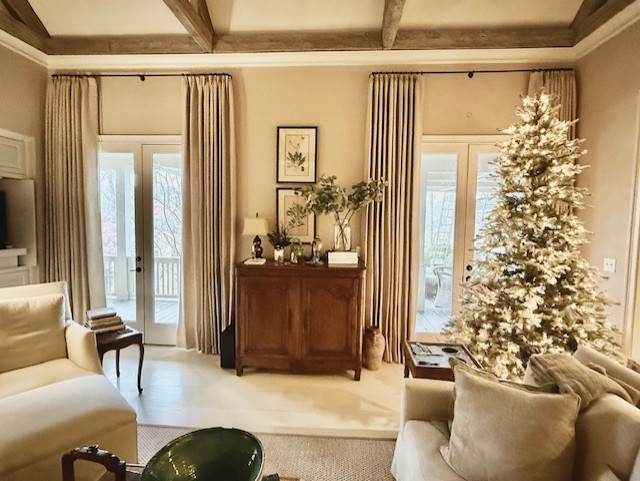 A cozy living room with cream furniture, two floor-to-ceiling windows with drapes, a wood cabinet, and a lit Christmas tree.