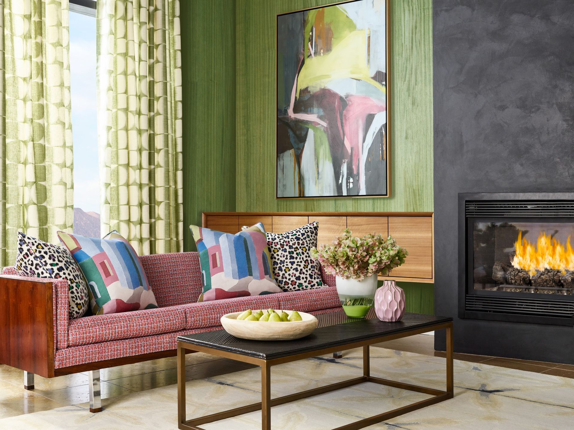A living room with a red patterned sofa, abstract artwork, green paneled walls, and a fireplace with a lit fire.
