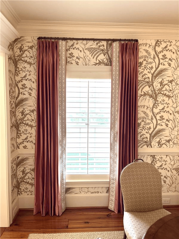 A window with rose-colored pleated curtains featuring decorative white trim, set against patterned floral wallpaper.