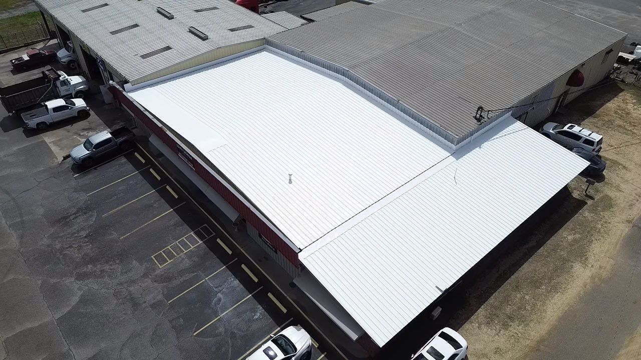 Aerial view of a commercial building with white metal roof and parking lot with several white vehicles.