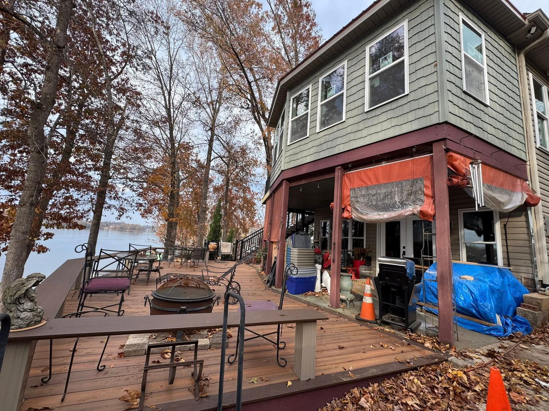 Lakeside house with deck. Brown leaves, tables, chairs. Deck partially covered with canopy. Lake view in background.