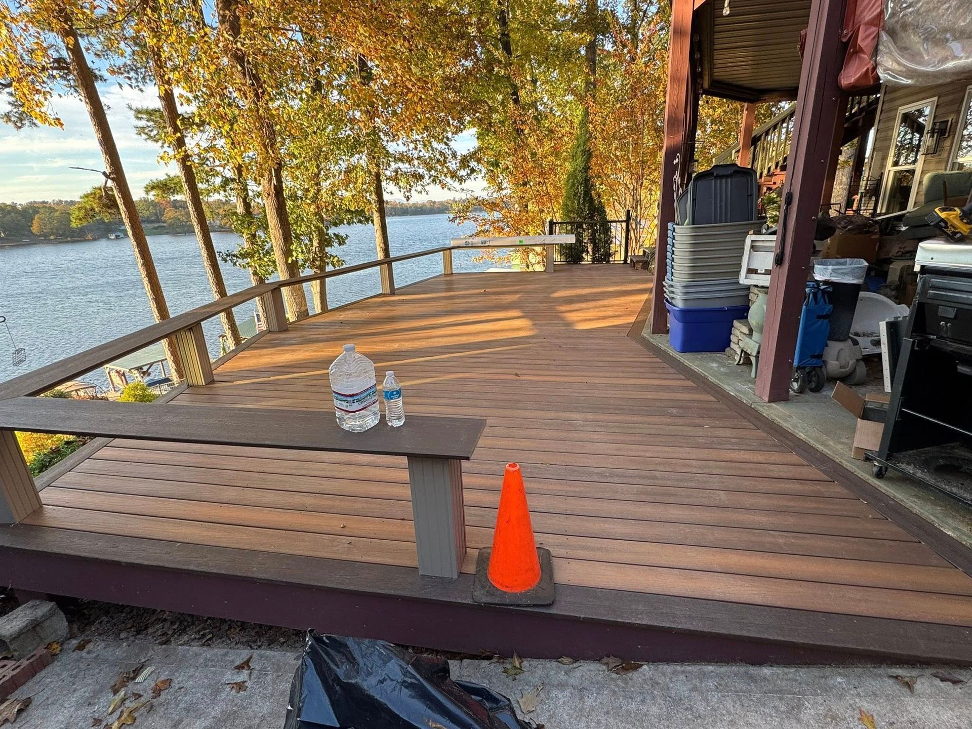 Wooden deck overlooking a lake, with a bench, orange cone, and water bottles. Fall foliage in the background.