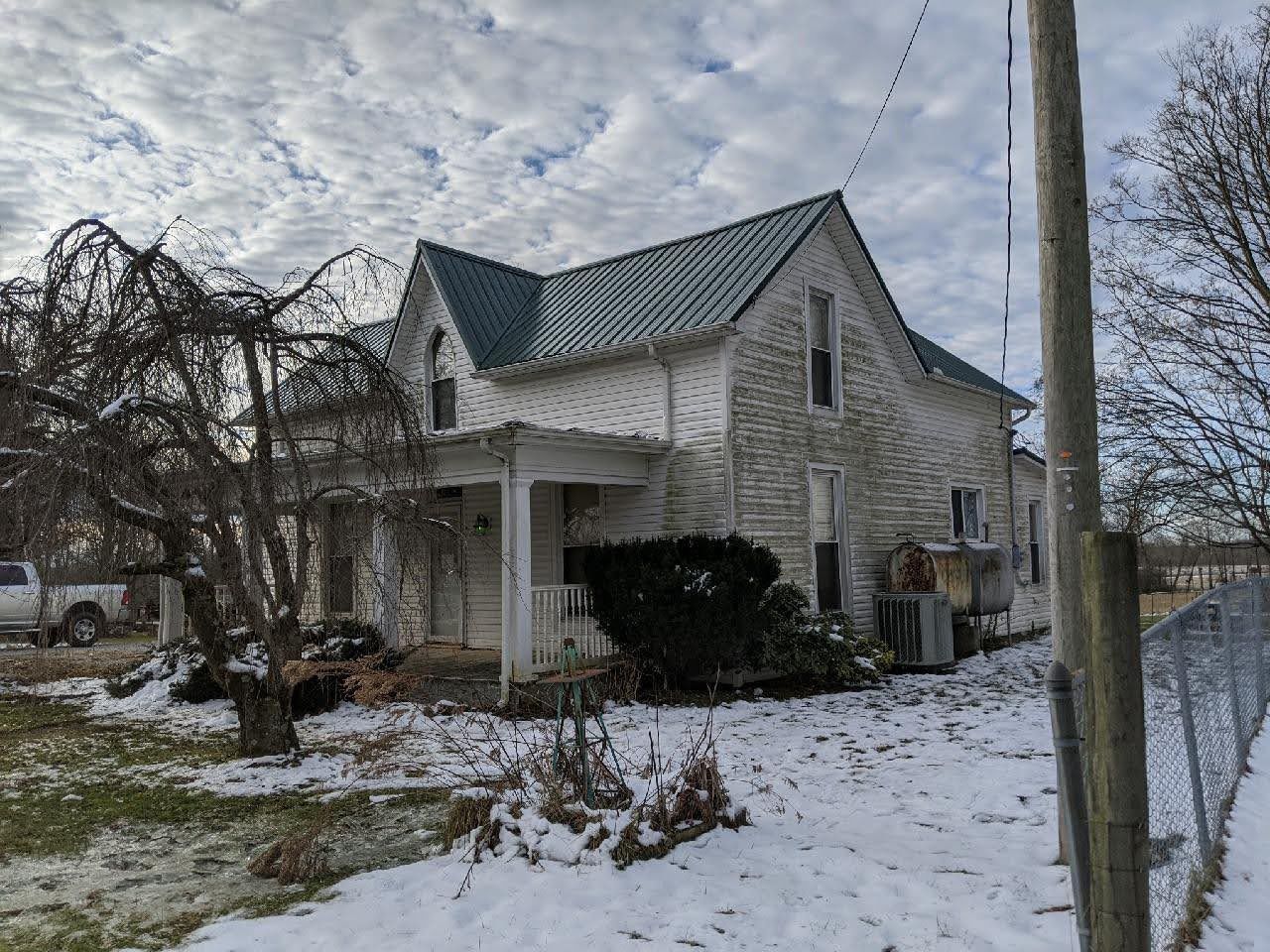 Old, weathered white house with green metal roof, snow-covered ground, cloudy sky.
