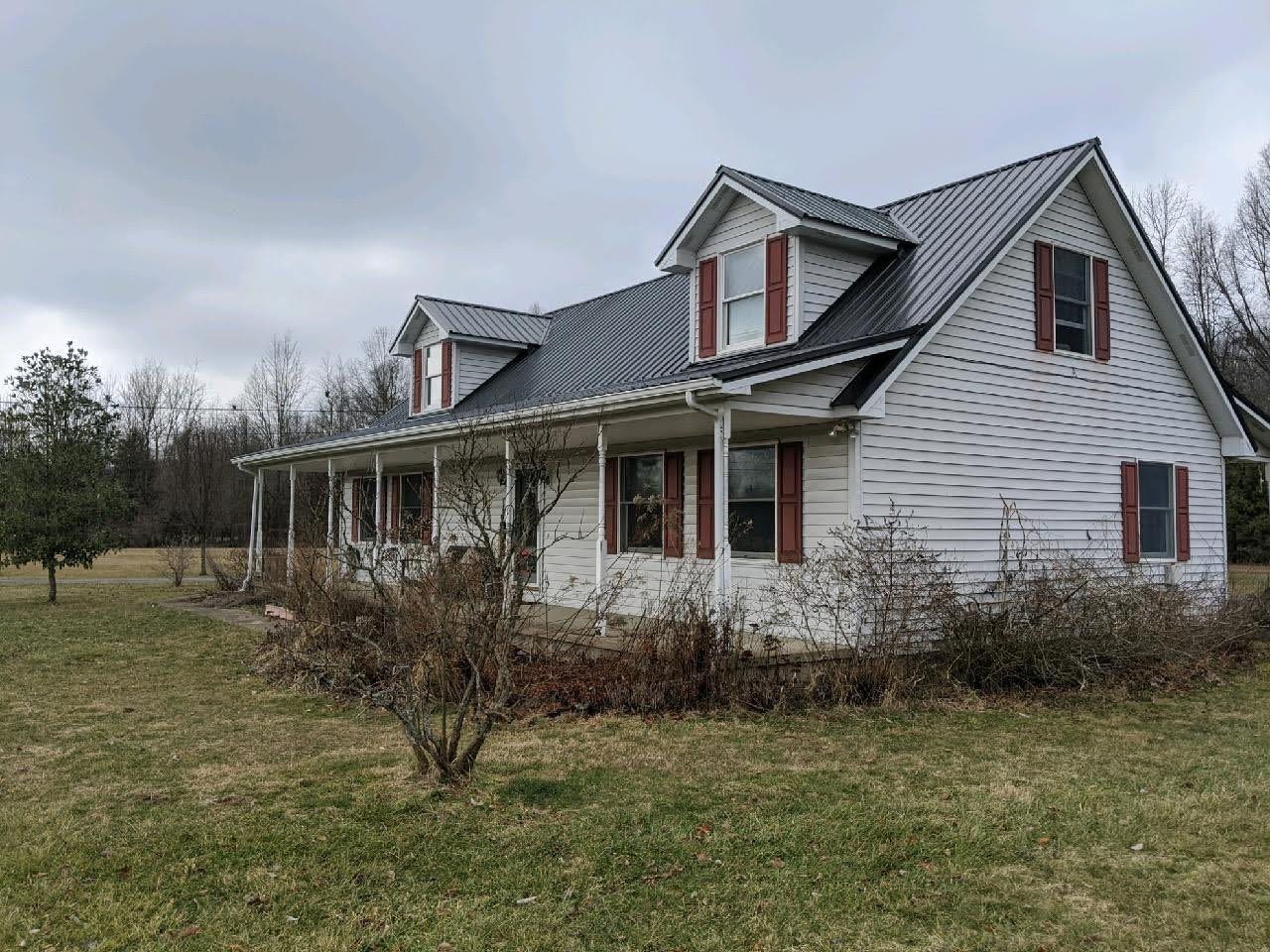White, two-story house with a wraparound porch and red shutters, under an overcast sky.