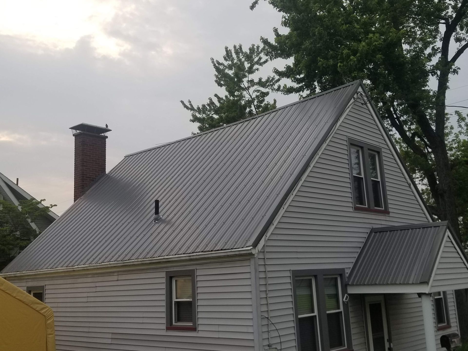 White house with gray corrugated metal roof, chimney, and porch roof. Overcast sky.