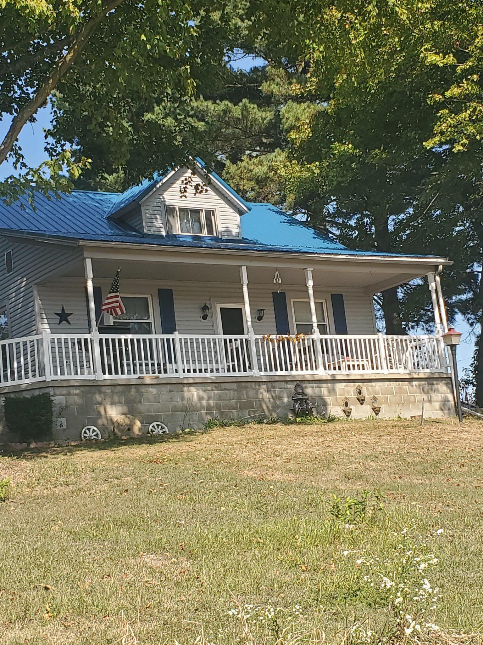 Cottage with blue roof, white porch and shutters, American flag. Overlooking a grassy hill with trees.