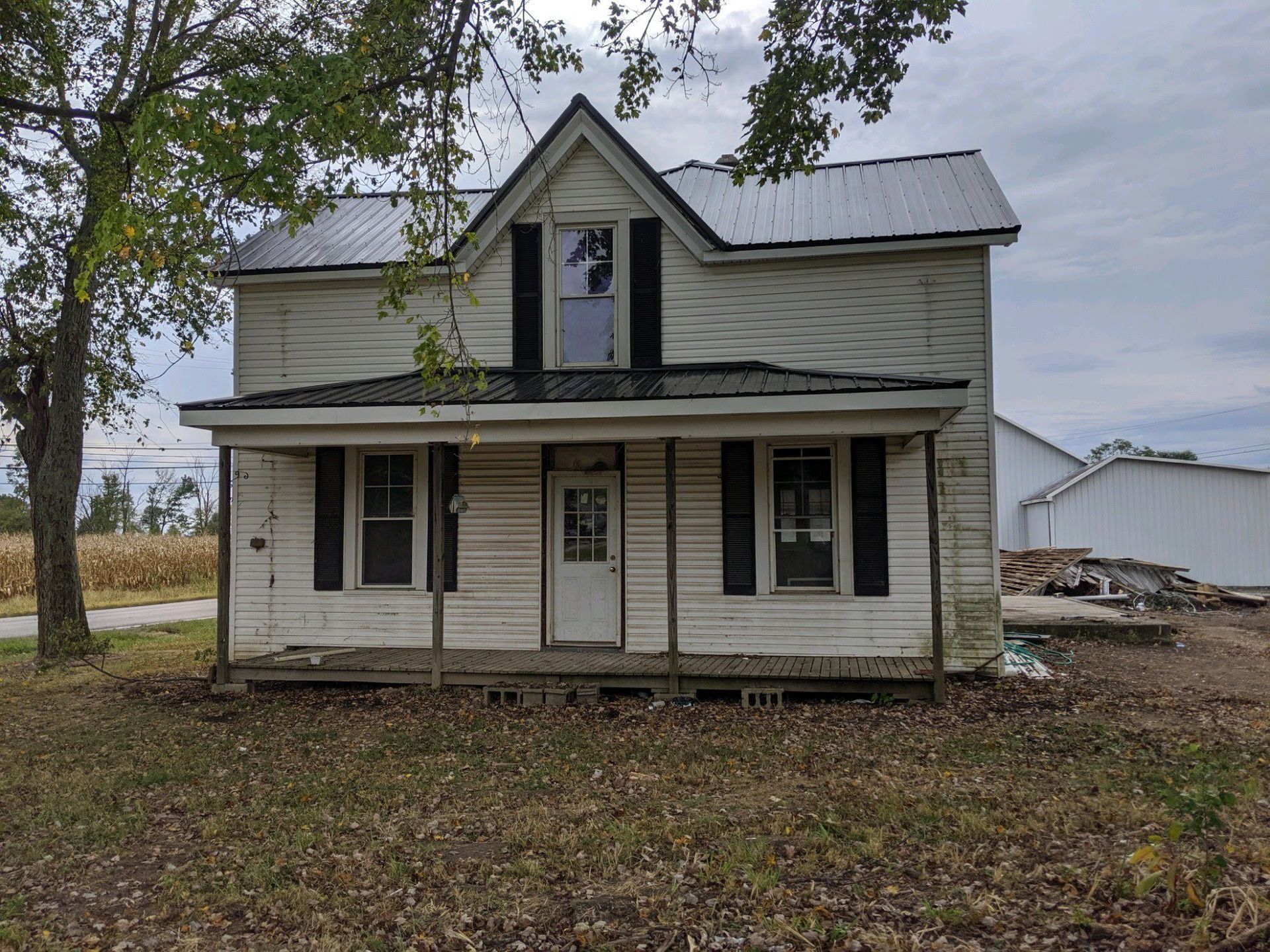 Dilapidated two-story farmhouse with peeling white paint, black shutters, and broken window. Overcast day.