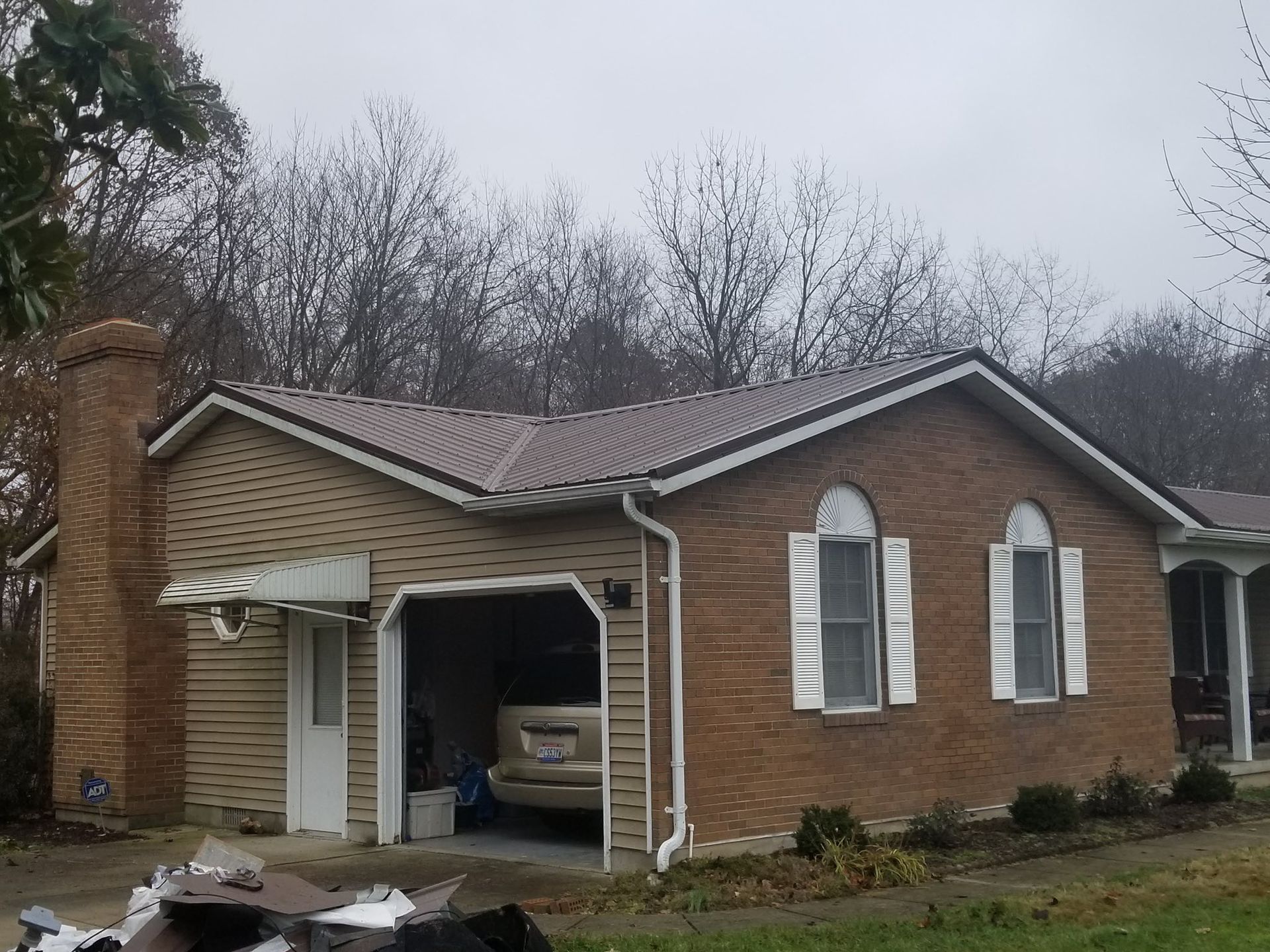 Brown brick and tan siding house with garage, chimney, and metal roof on a cloudy day.