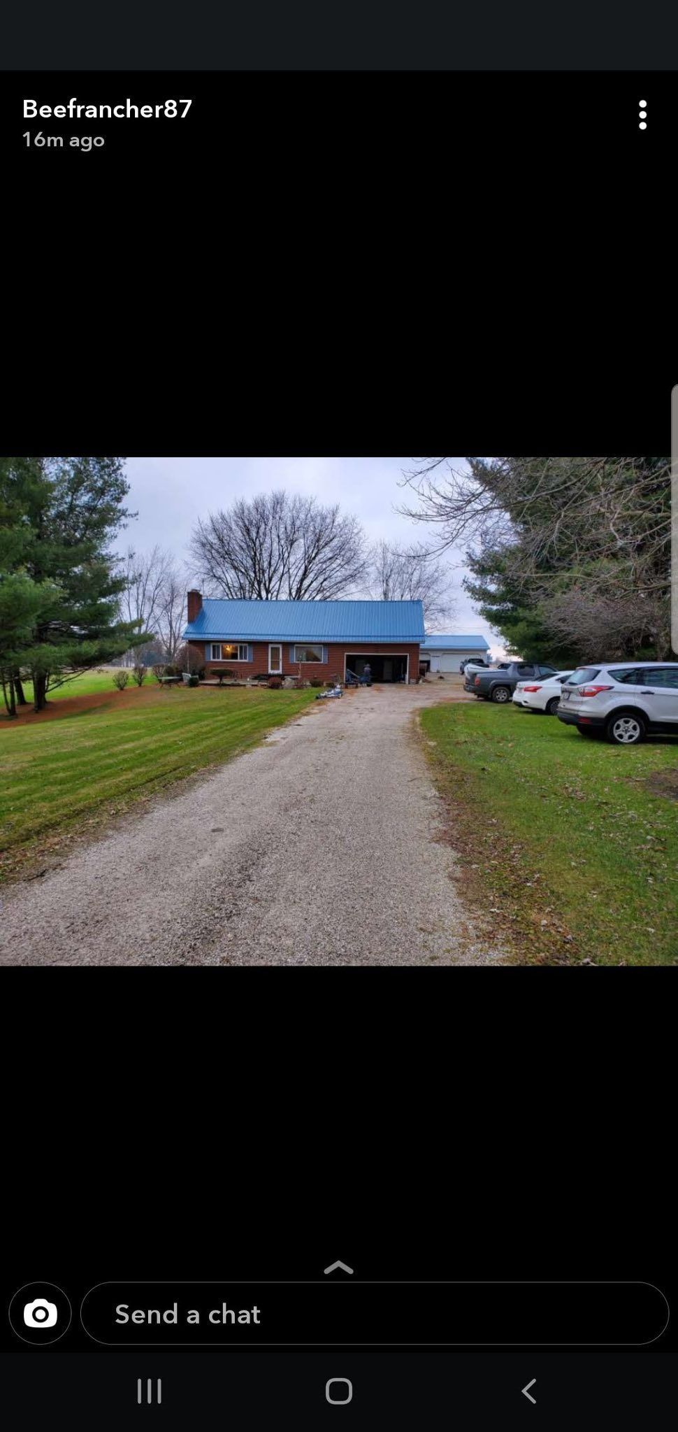 Gravel driveway leading to a small house with a blue roof. Cars parked nearby. Overcast sky.