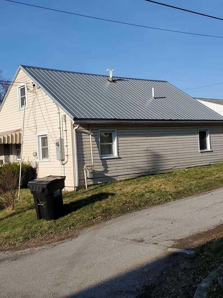 Tan house with a silver metal roof, black trash can, and a sloped driveway.