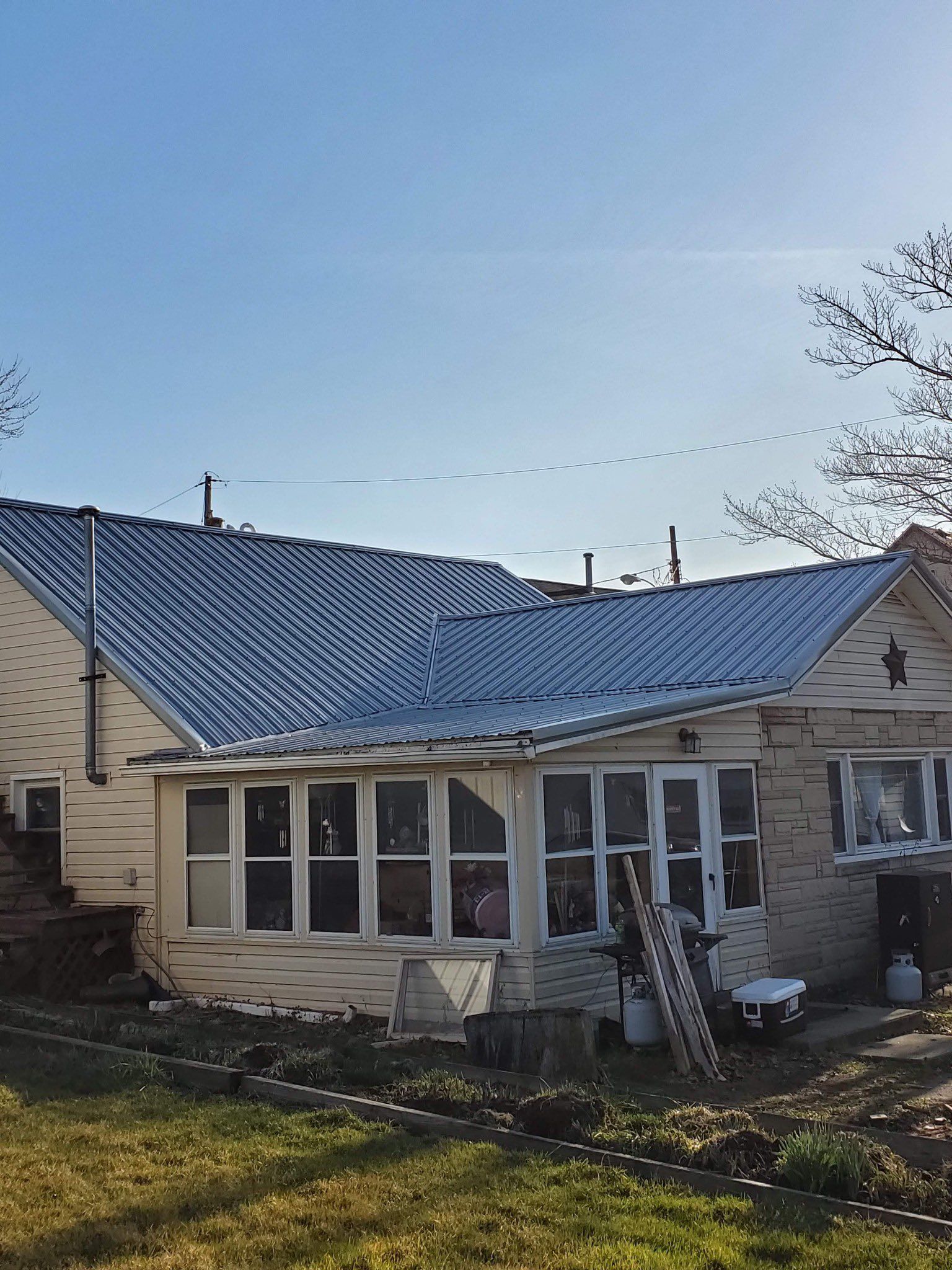 Off-white house with a corrugated metal roof, sunny day. Windows in a porch area, ladder leaning against the side.