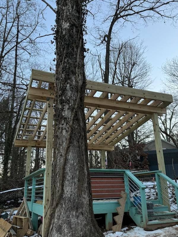 Wooden deck with pergola under construction, built around a tree. Teal deck, wood framing.