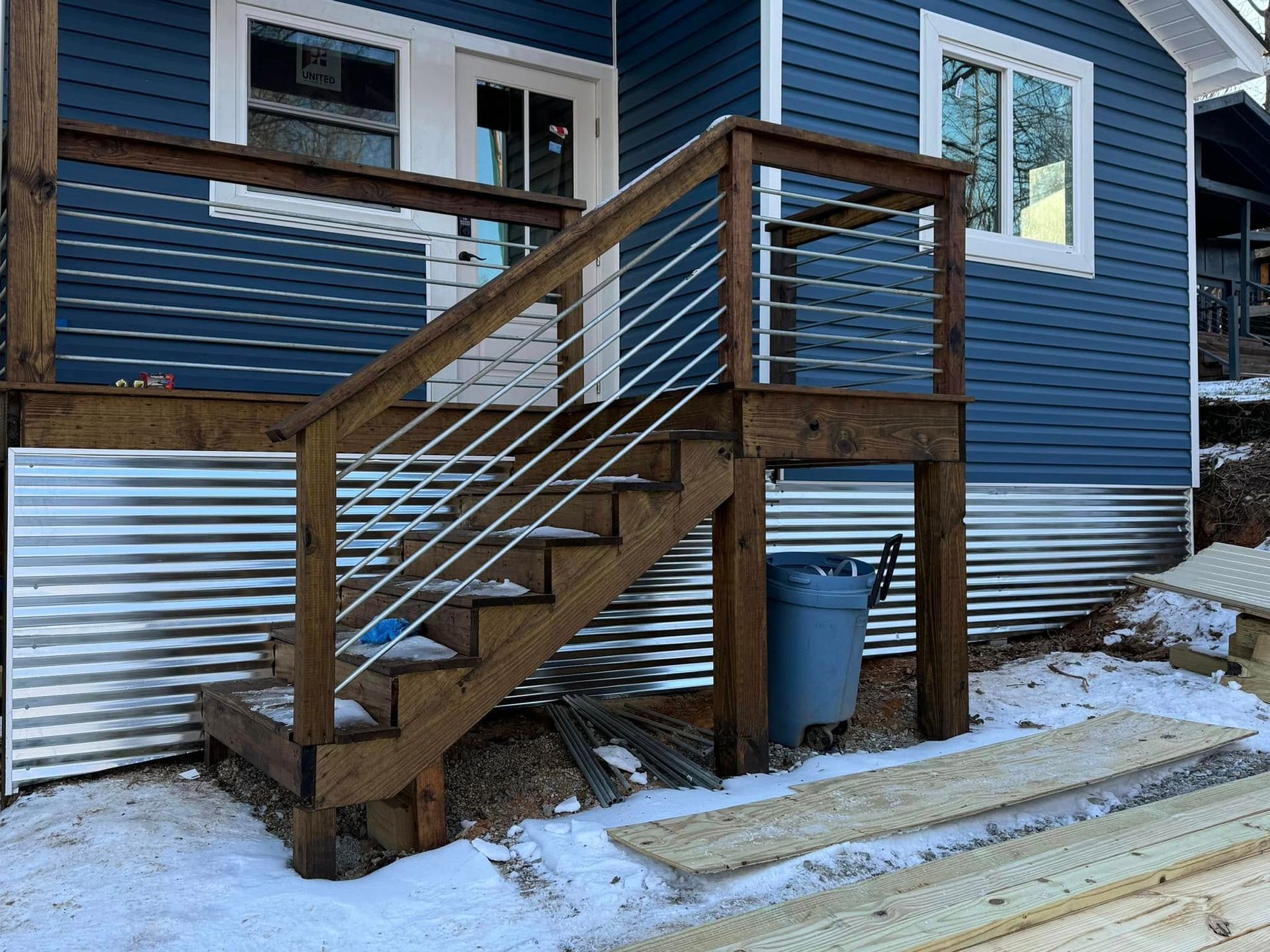 Wooden staircase with metal railing leading to a blue house with corrugated metal siding. Snow on the ground.
