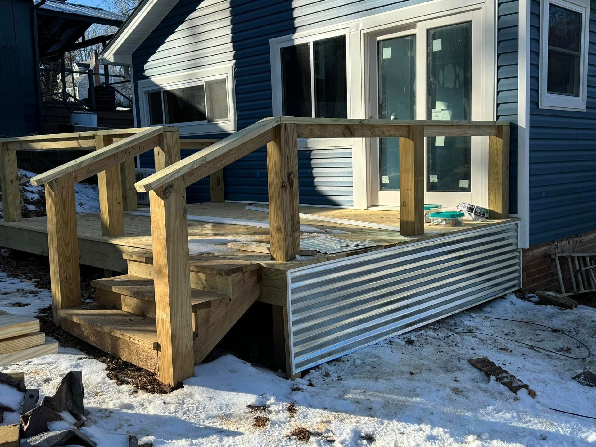 Wooden deck with stairs, handrails, and corrugated metal skirting against a blue house with sliding glass doors.