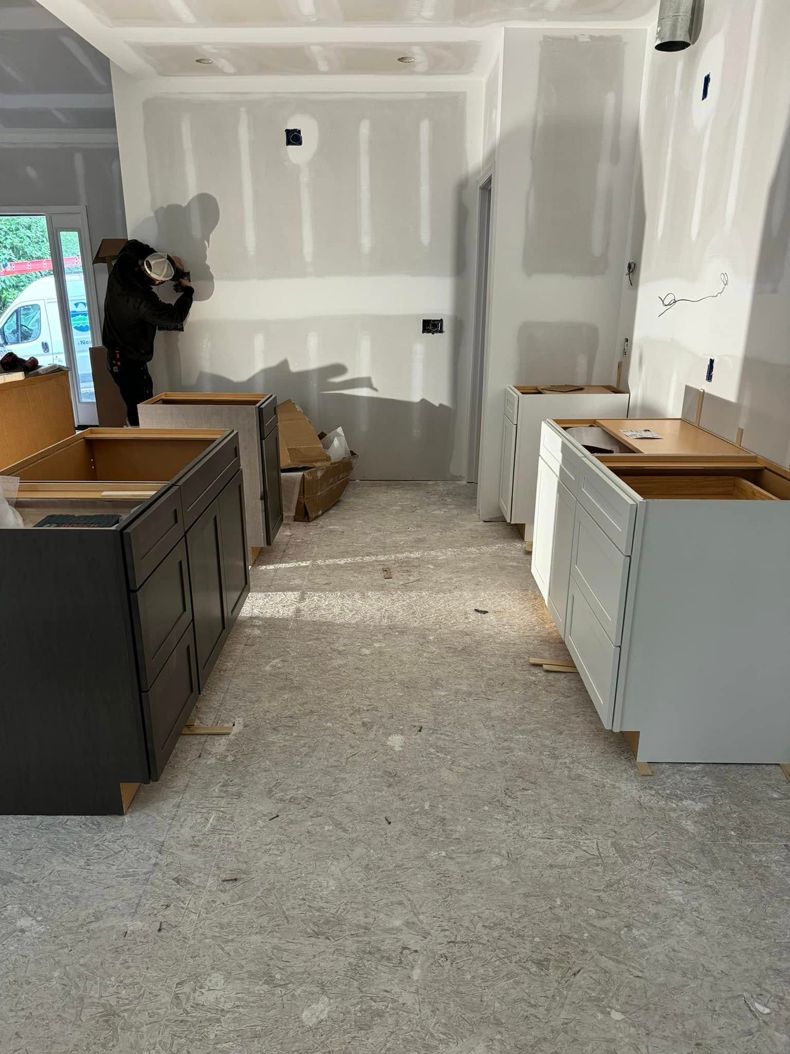 Kitchen under construction: cabinets in gray and white, drywall, worker in the background.