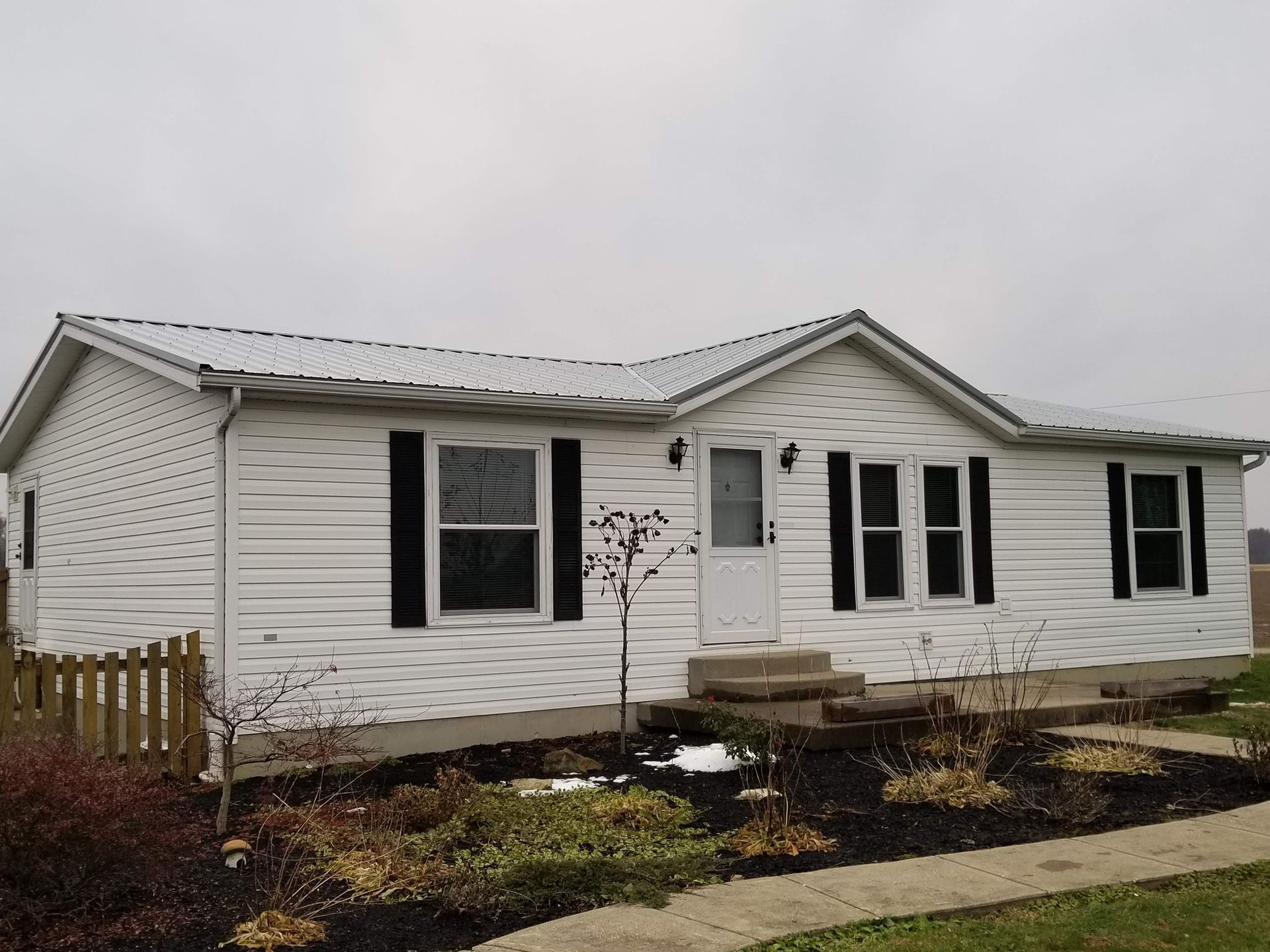 White house with black shutters, metal roof, and small front yard with dark mulch and some snow.
