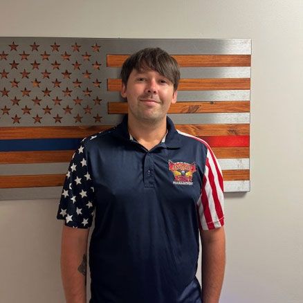 Man wearing a polo shirt with American flag design, standing in front of an American flag wall hanging with a blue and red stripe.