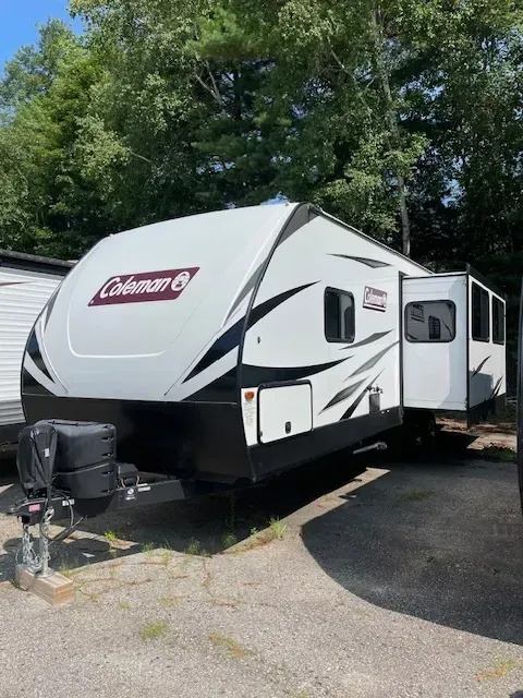 A white and black coleman trailer is parked in a gravel lot.