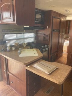 RV kitchen with brown cabinets, countertops, and appliances.  View includes oven, microwave, refrigerator, and window.