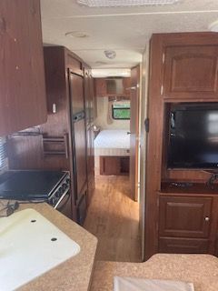 RV interior: kitchen with stove and sink, hallway leading to bedroom with bed and TV on cabinet.