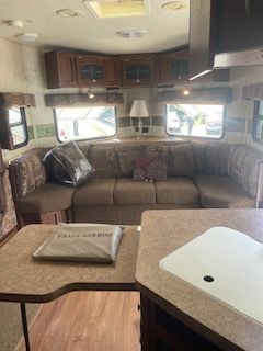 Interior of a travel trailer: brown sectional sofa, kitchen counter, upper cabinets, and windows.