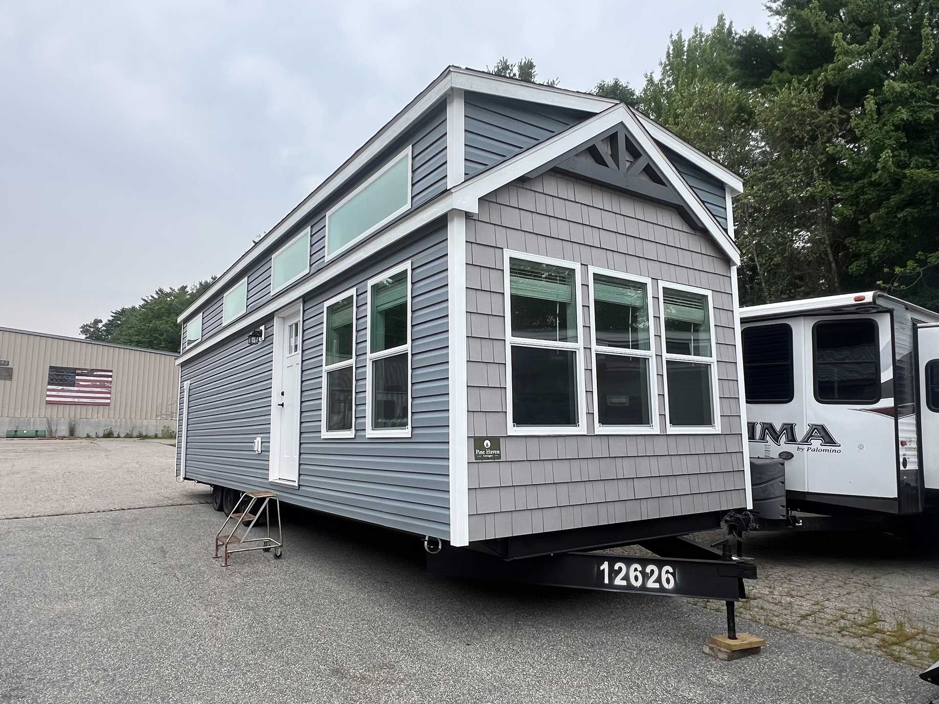 tiny house on wheels with gray and blue siding, many windows, parked on gravel