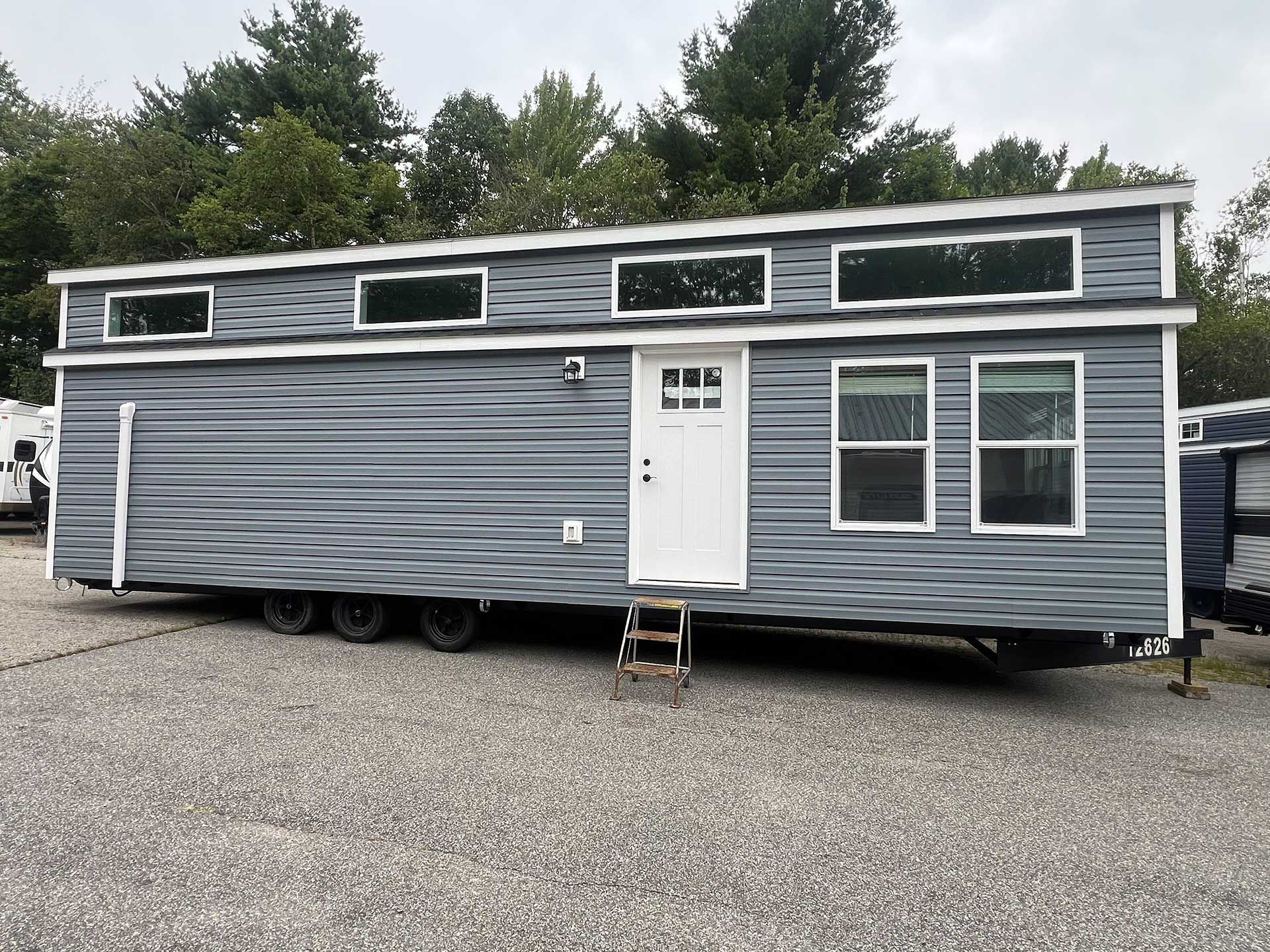 gray tiny house on trailer, white door, windows, gravel lot, trees in background