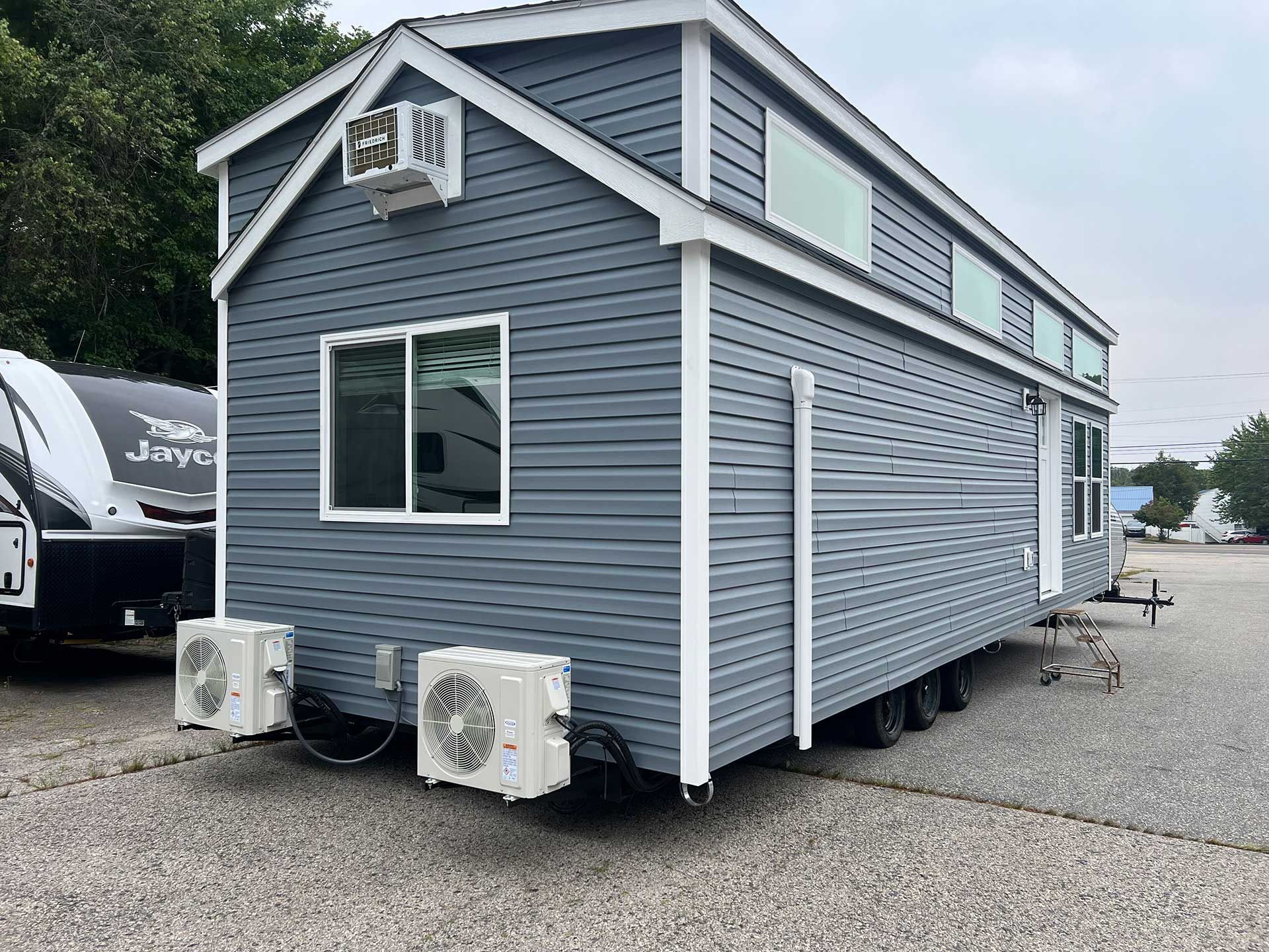 gray tiny home on wheels with air conditioning units, parked on gravel