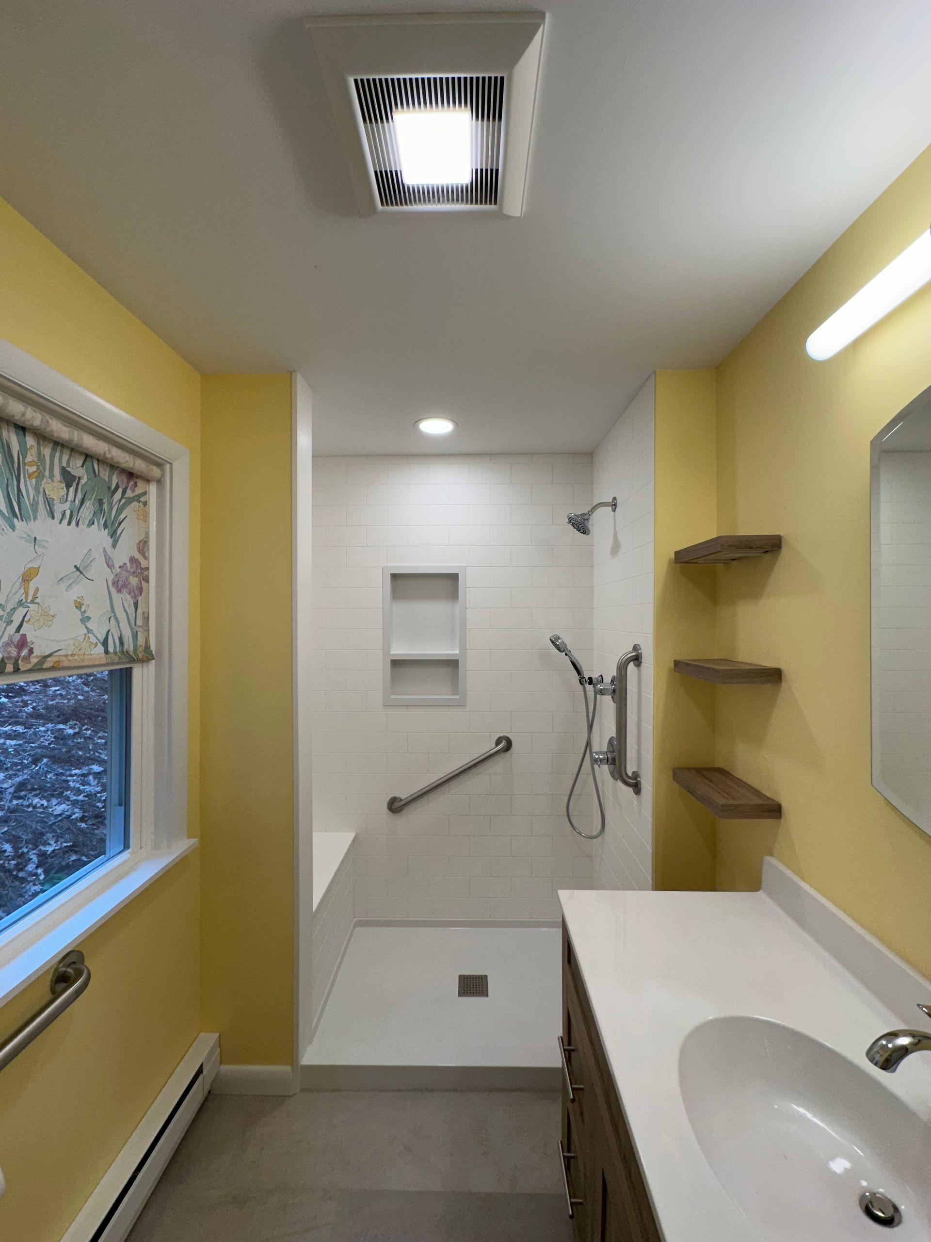 Yellow bathroom with white shower, shelves, and vanity. Window with floral shade. Ceiling fan.