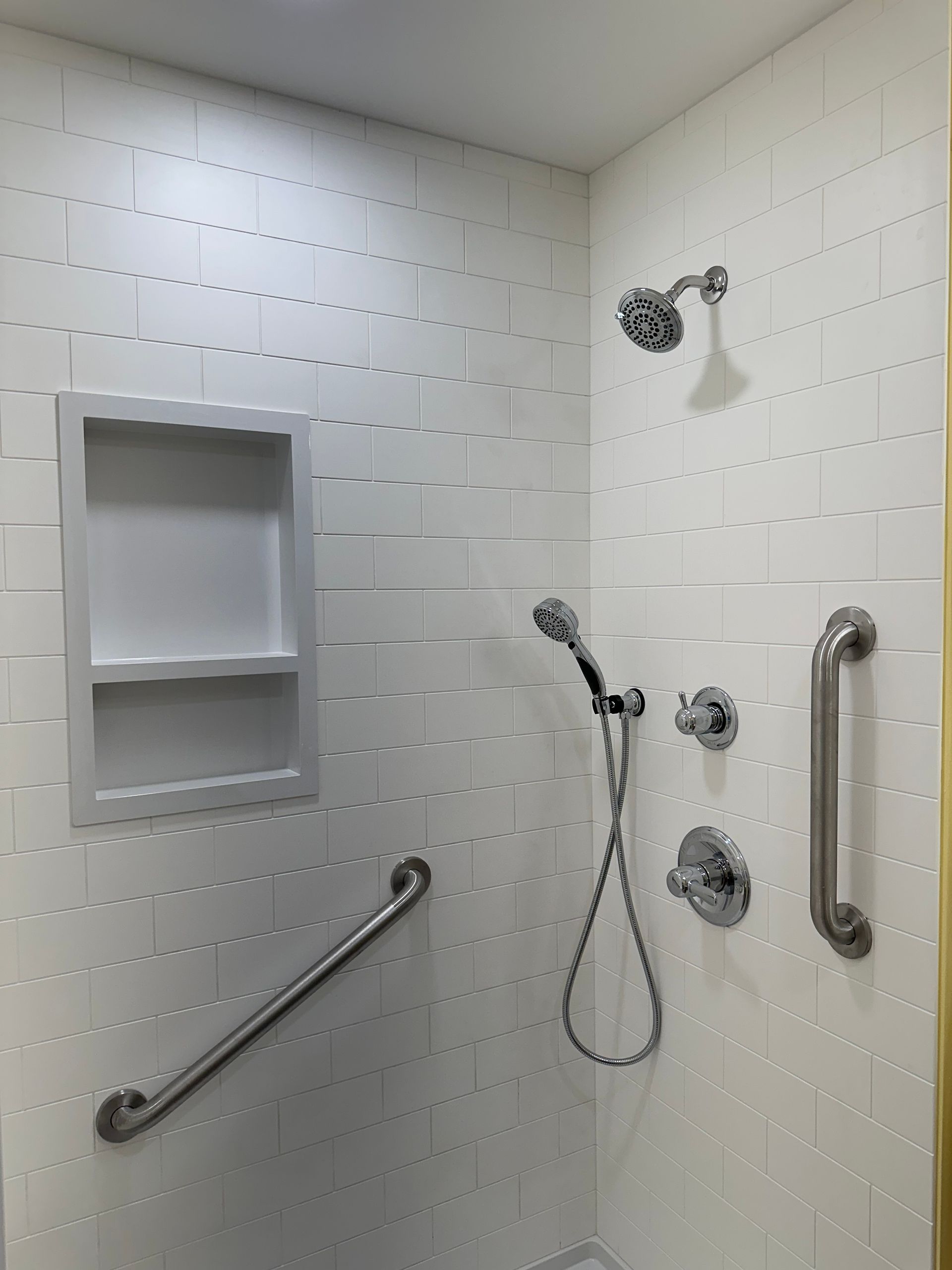 White tiled shower with grab bars and built in shelf. Silver shower head and handheld shower.