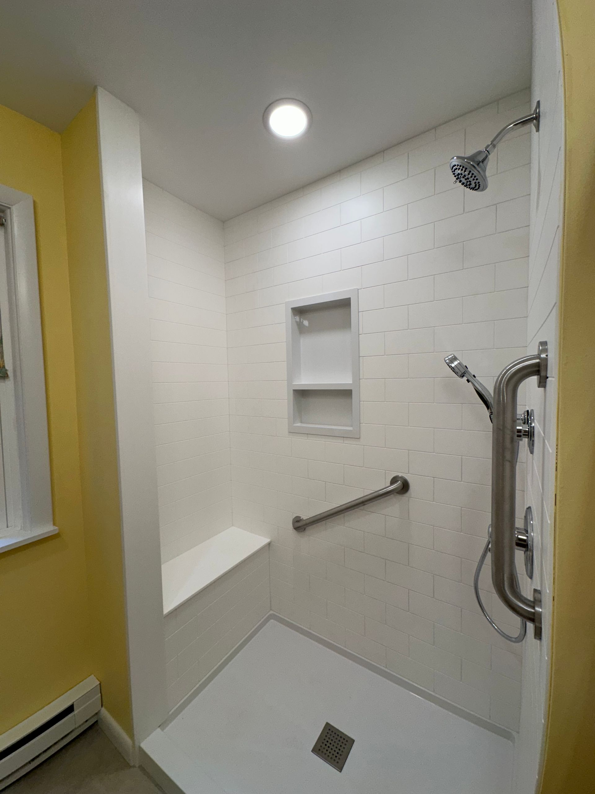 White tiled shower with built in seat, grab bar, and recessed shelf.