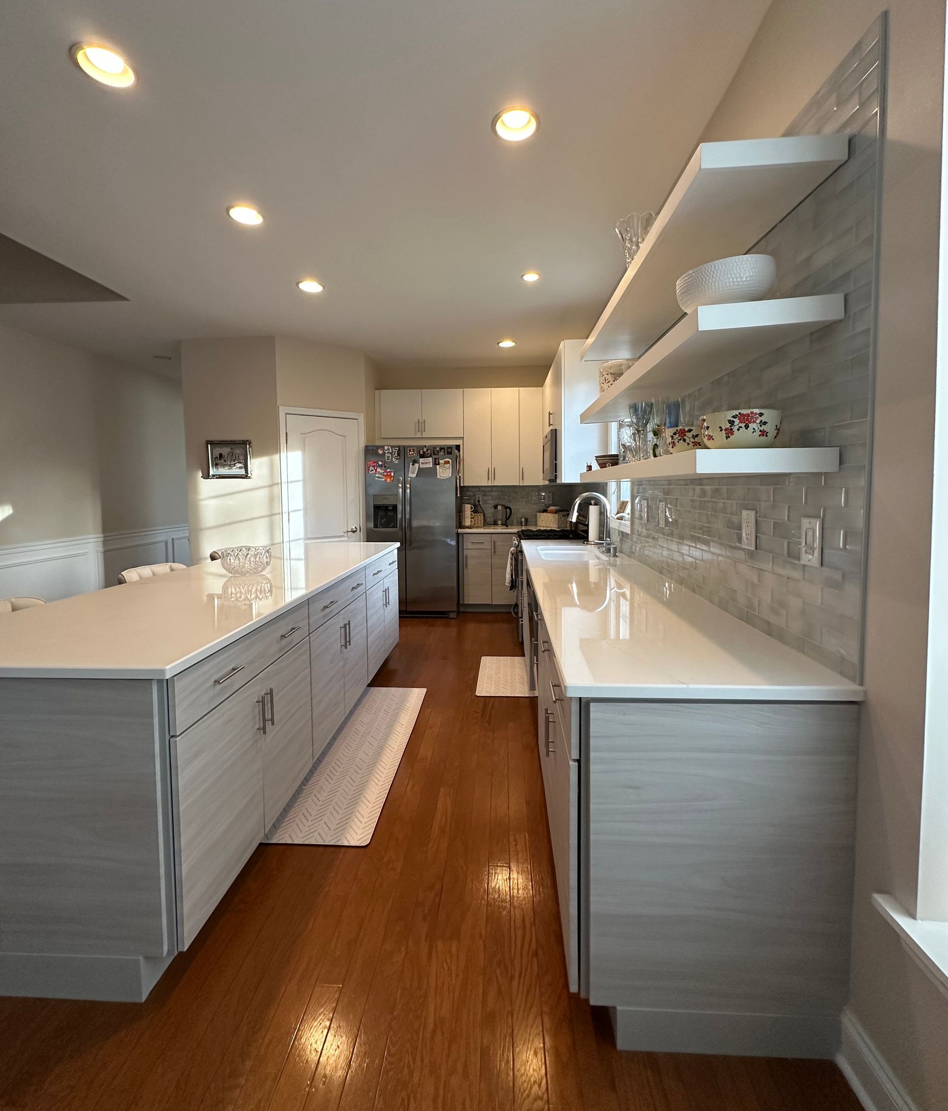 Modern kitchen with light gray cabinets, white countertops, and wooden floors. Open shelving and a large island.