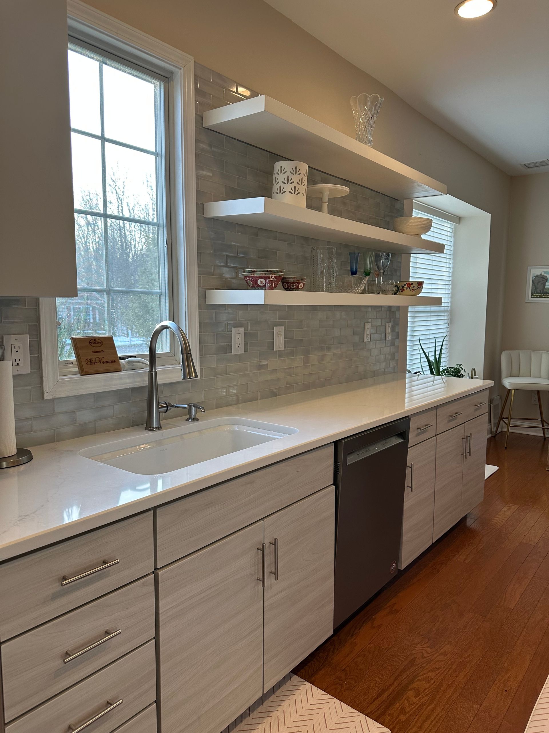 Modern kitchen with white cabinets, countertops, and backsplash, a stainless steel sink and dishwasher, and floating shelves.