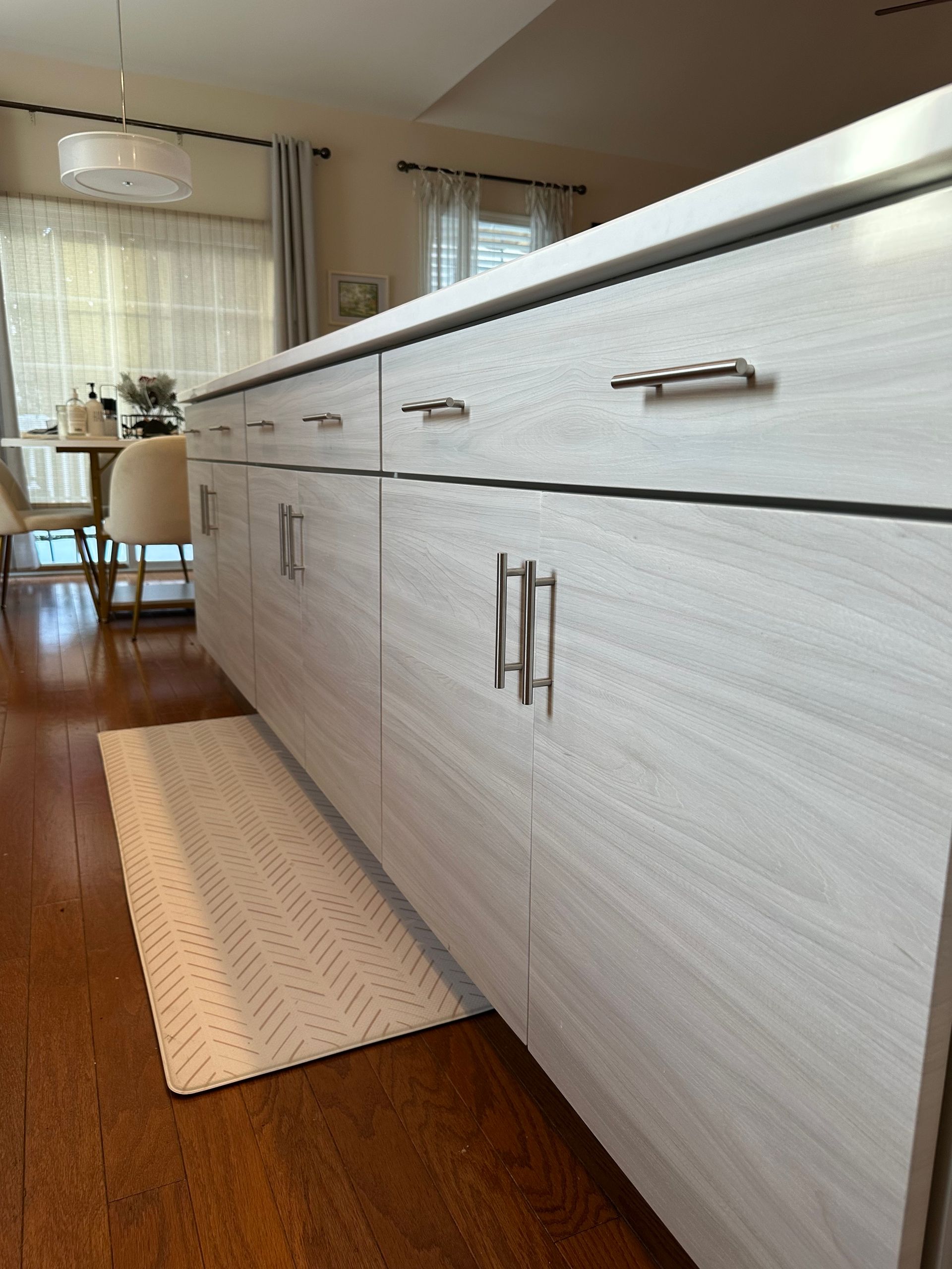 Kitchen island with light colored cabinets and countertops, stainless steel hardware, and a small rug on a hardwood floor.