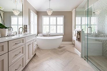 A bright beige bathroom with a white soaking tub, large window with shutters, glass shower, and light wood herringbone floor