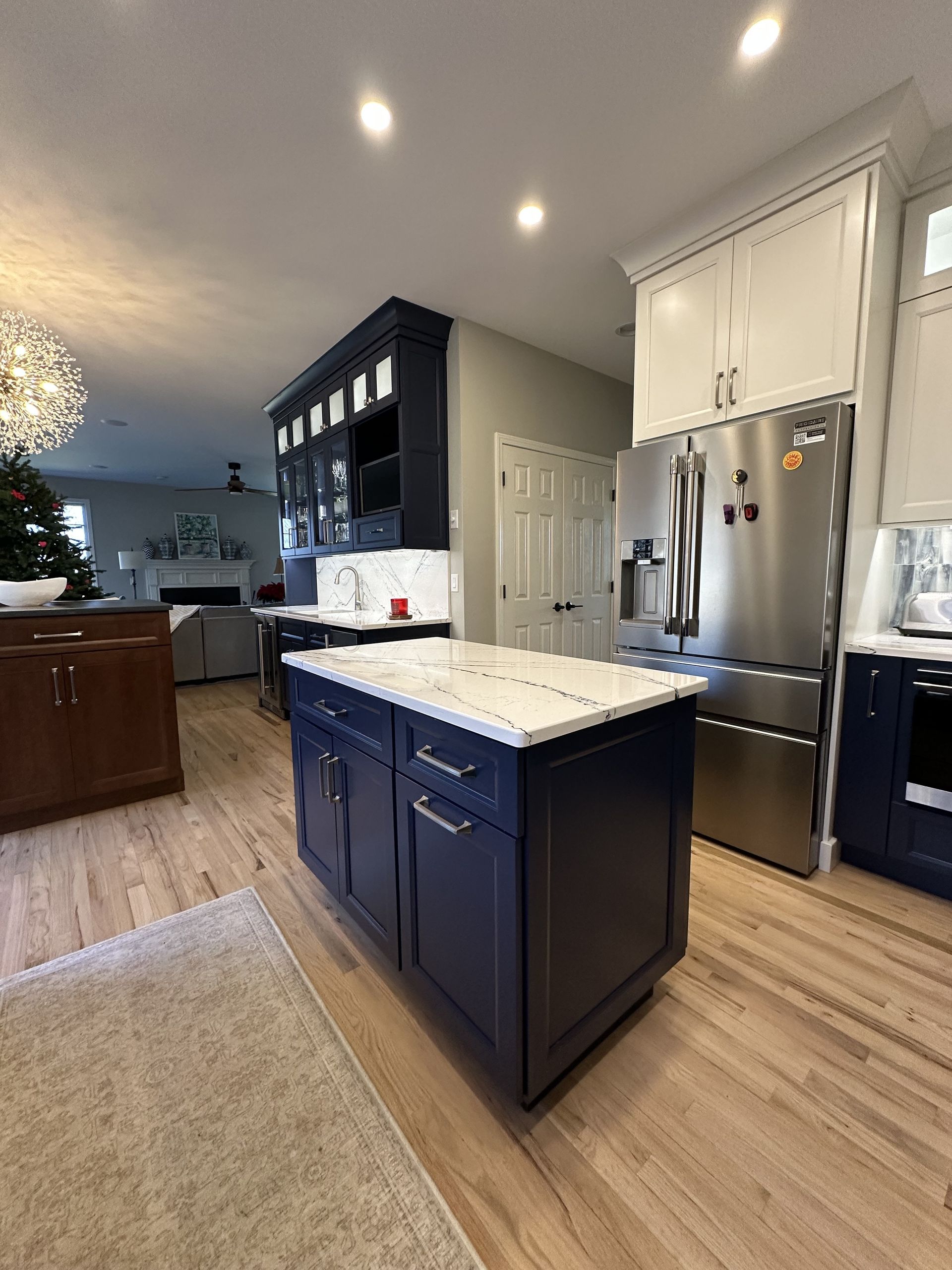 Blue and white kitchen with island, stainless steel fridge, light wood floors, and recessed lighting.