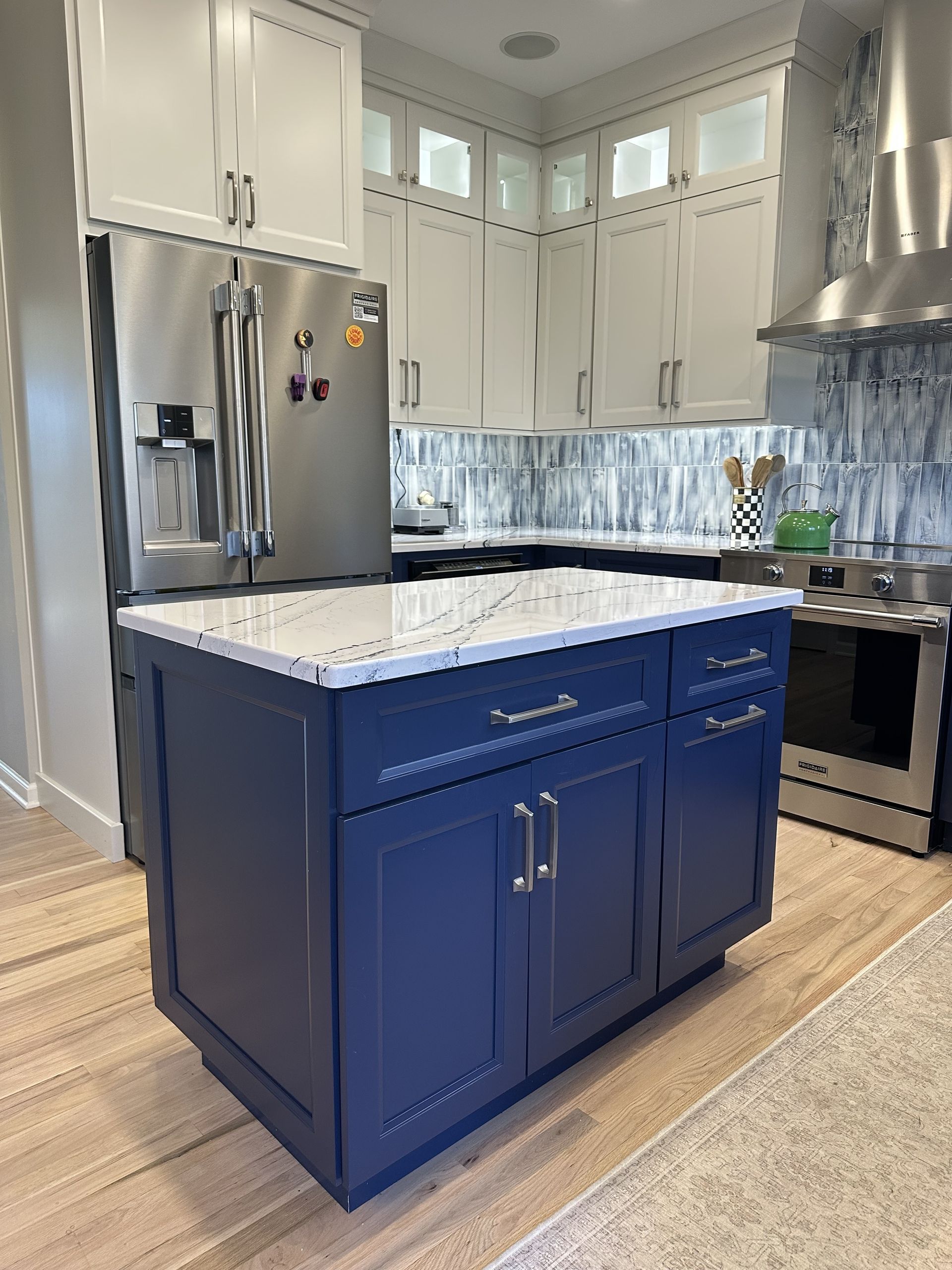 Blue kitchen island with white countertop, white and blue cabinets, stainless steel appliances, and a light-colored wood floor.