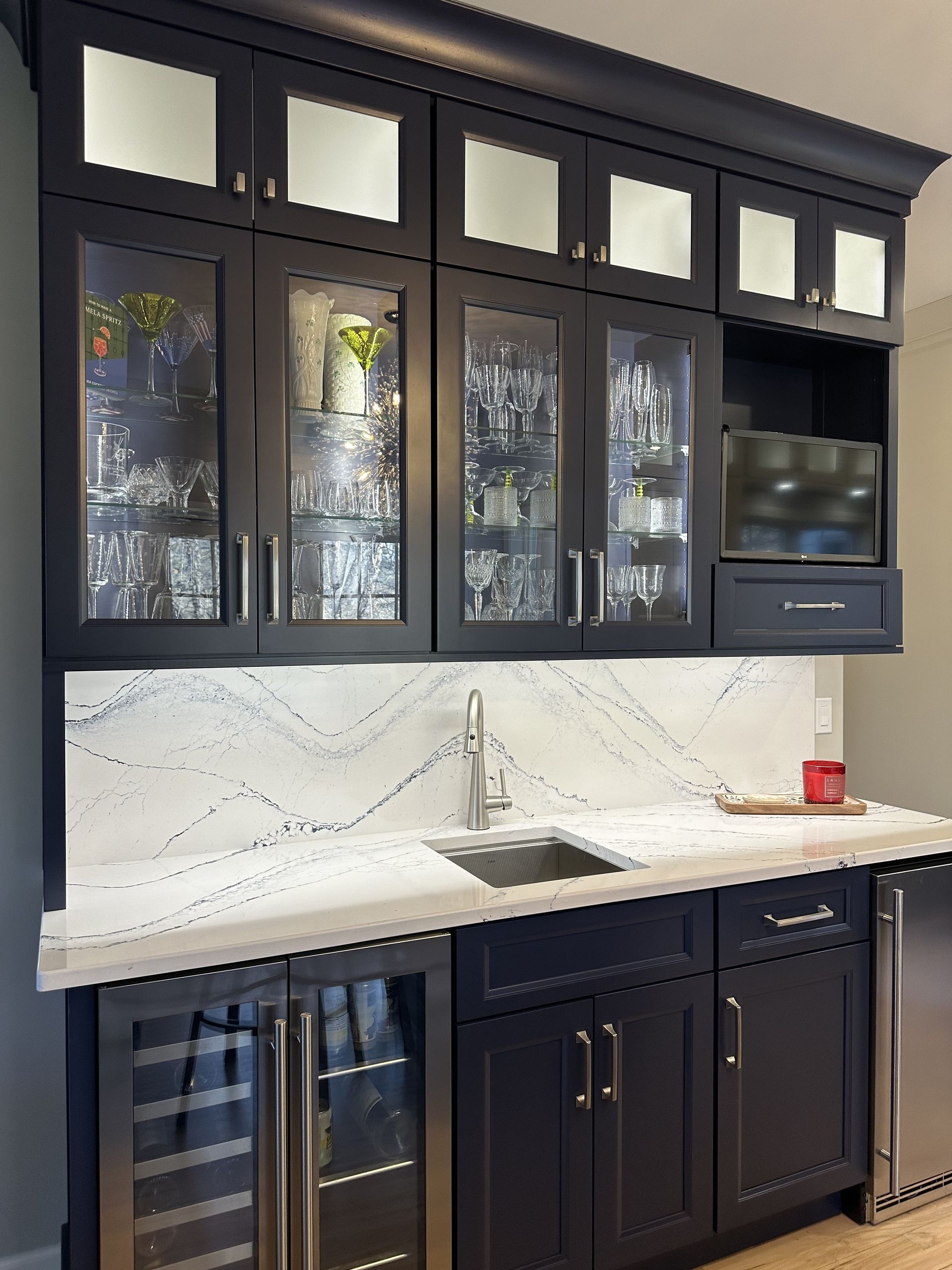 Dark blue home bar with white countertop, sink, wine fridge, and cabinets. Glass-front display cabinets.