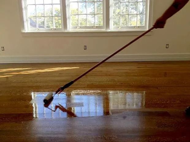 Person using a long-handled roller to apply wood finish to a hardwood floor in a room with windows.