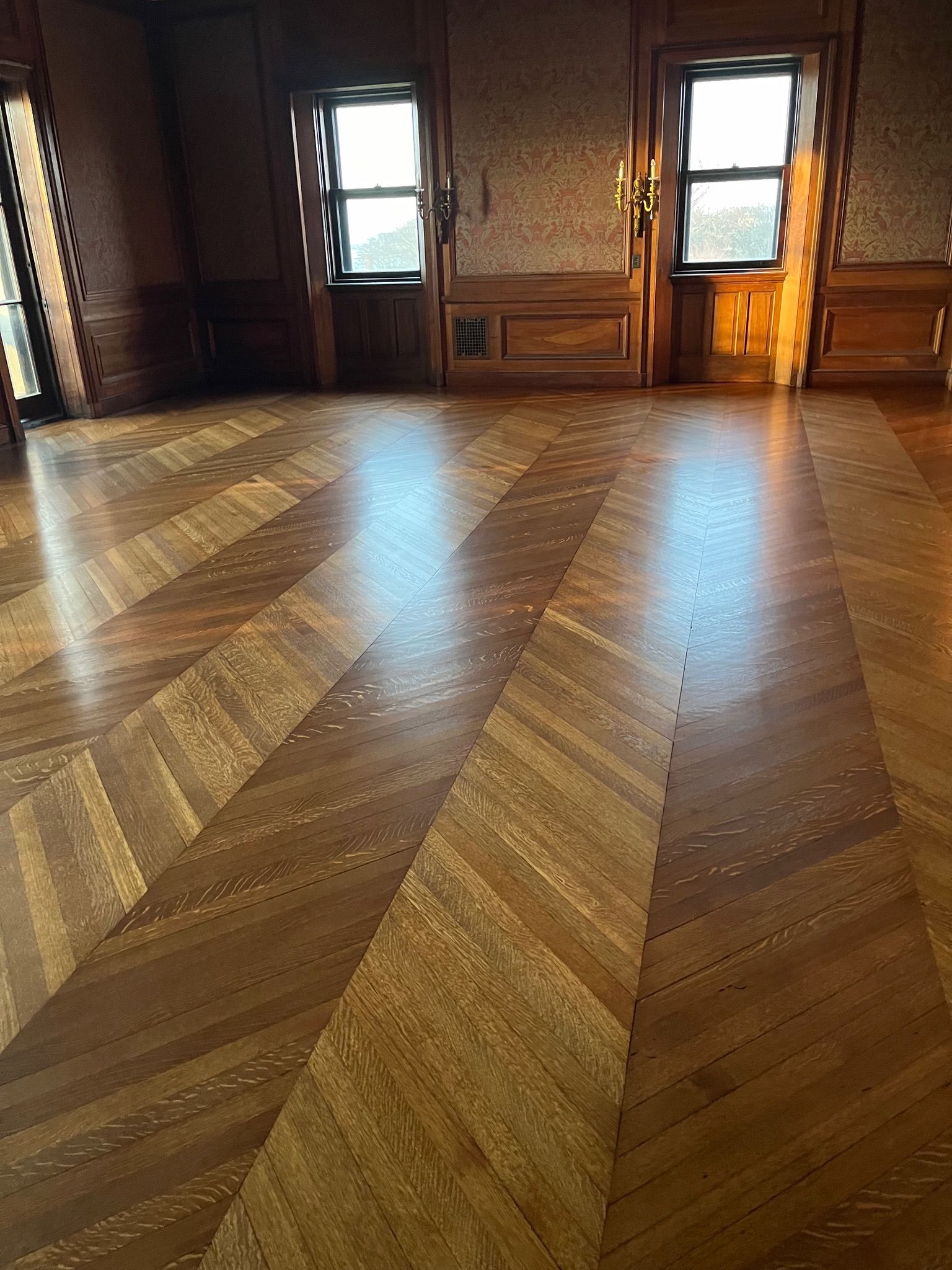 Wooden parquet floor with a chevron pattern. Two windows and wood paneling are in the background.
