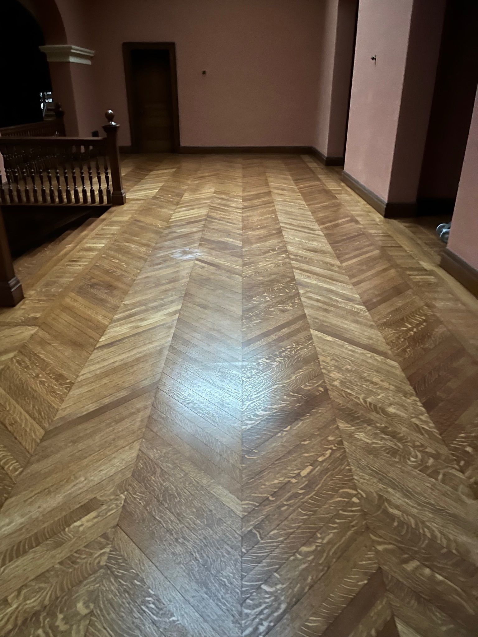 Herringbone pattern wood floor in a long hallway with pink walls and a wooden banister on the left.