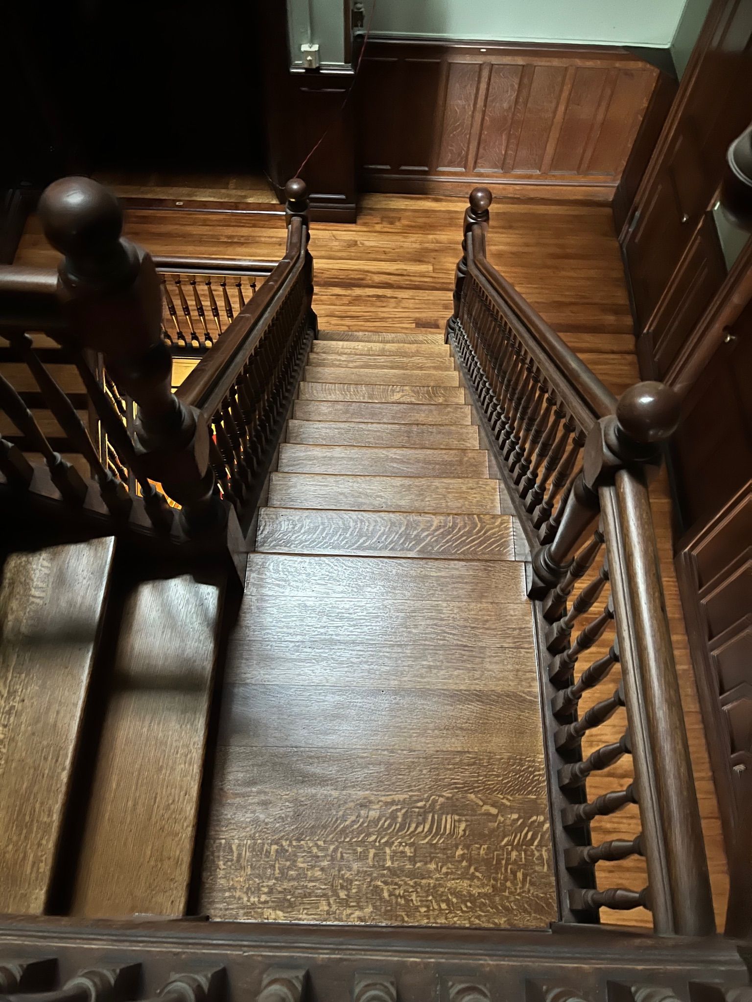 Wooden staircase with dark railing and balusters, leading down from a hallway with wooden paneling.