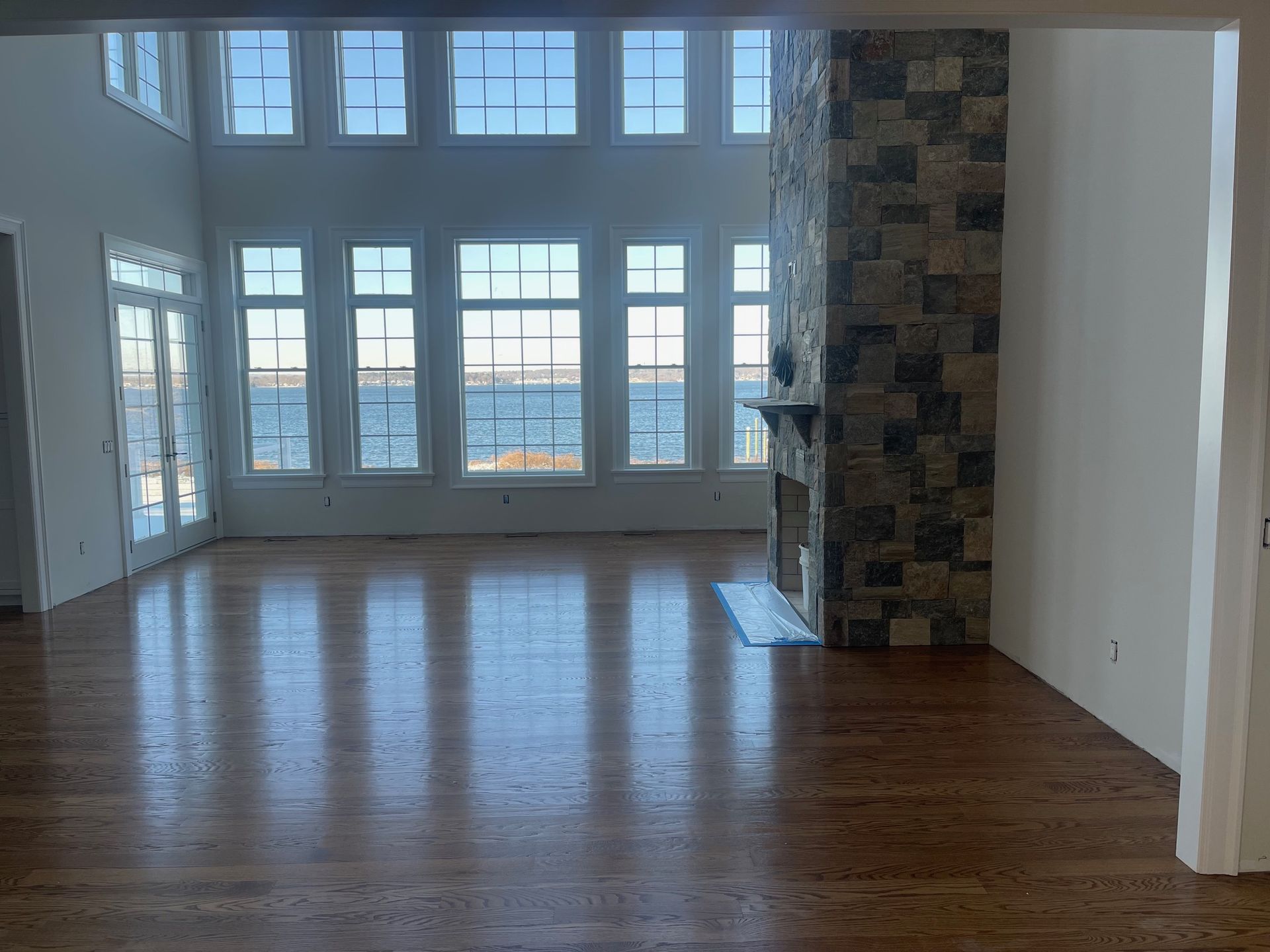 Spacious room with hardwood floors, large windows overlooking water, and a stone fireplace.